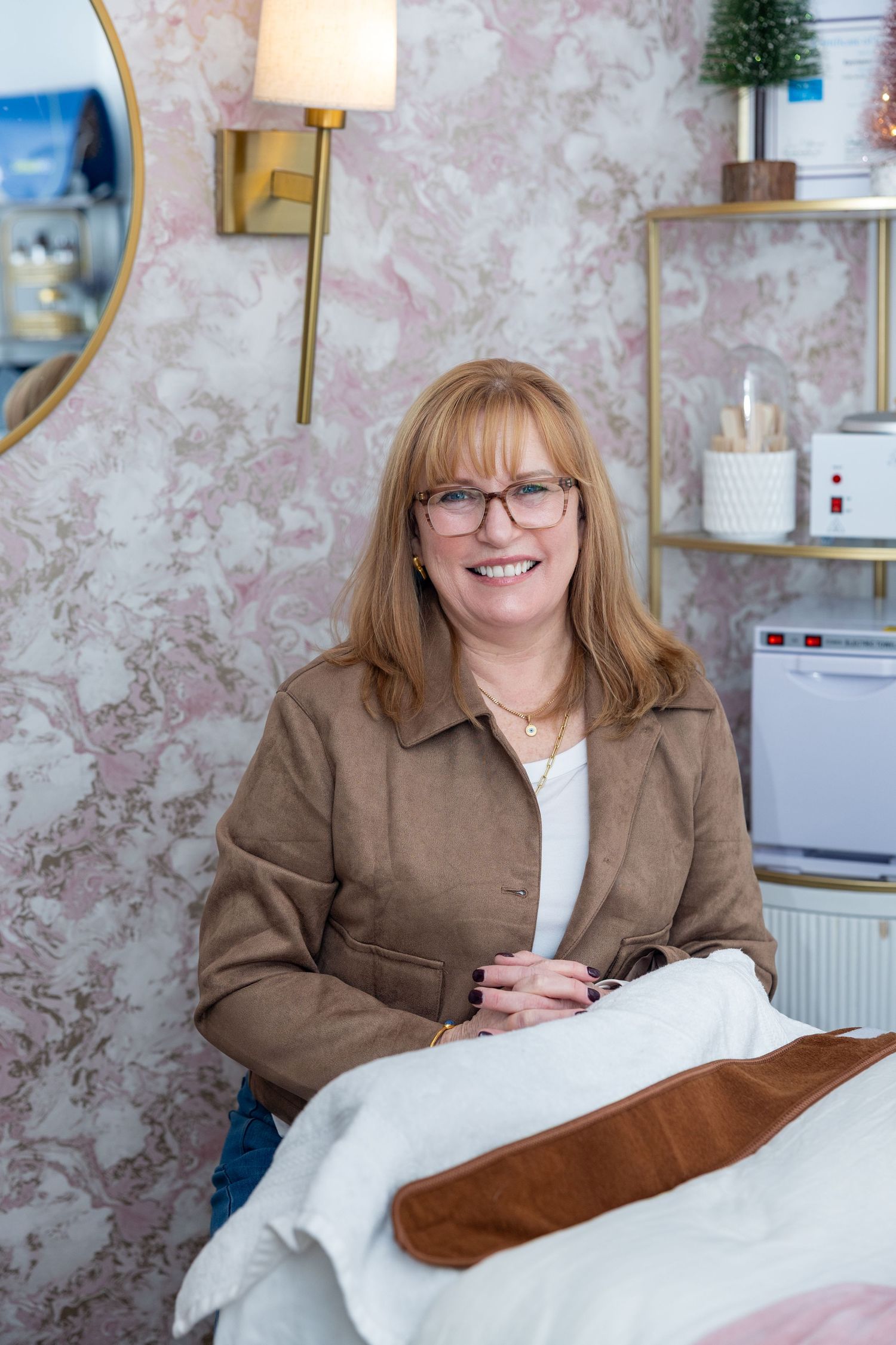 Esthetician wearing glasses and brown blazer poses in her spa for professional business headshot photos. Spa bed and equipment included.