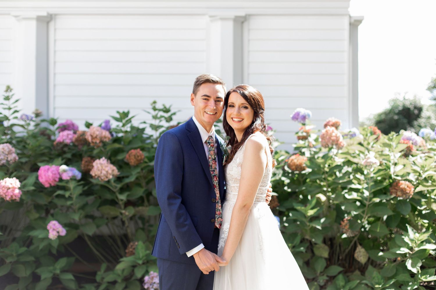 Smiling bride and groom posing together in front of blooming hydrangea bushes at an outdoor wedding venue.