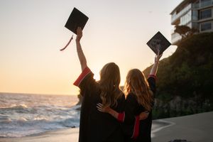 Two graduates raise caps toward ocean horizon at sunset on a scenic California beach.