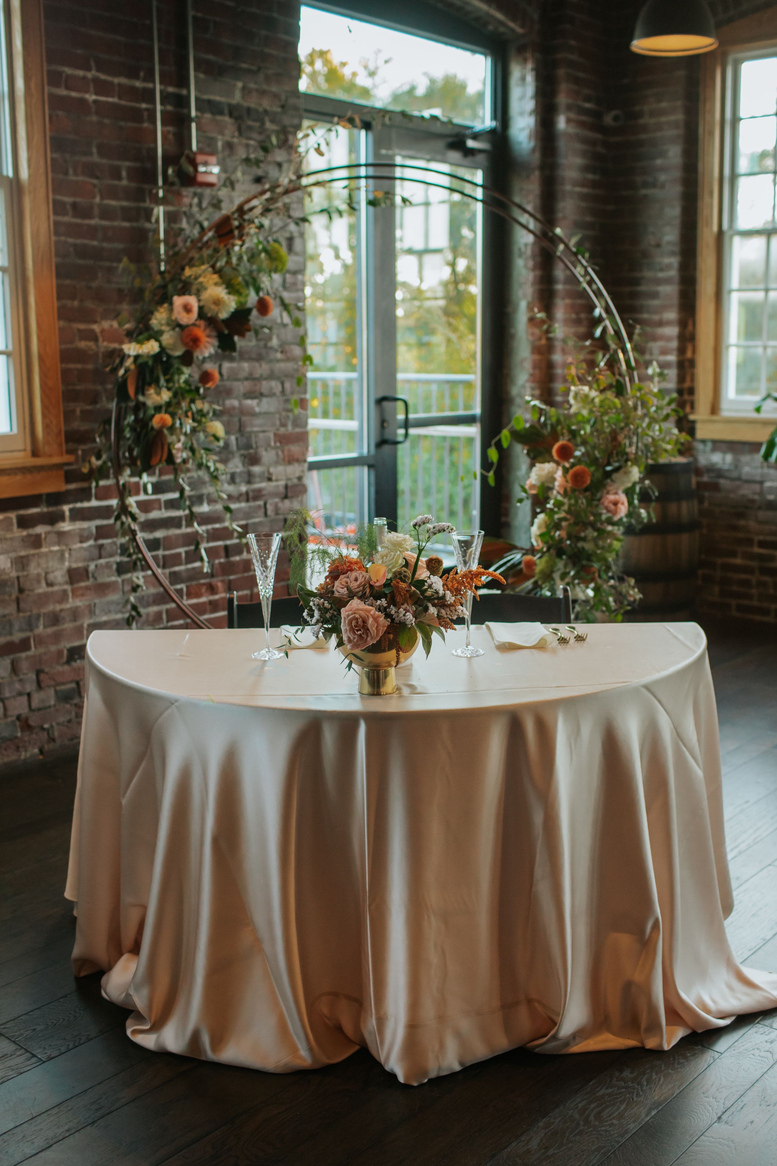 Elegant round tables with champagne-colored satin tablecloths and floral centerpieces in an industrial brick venue.