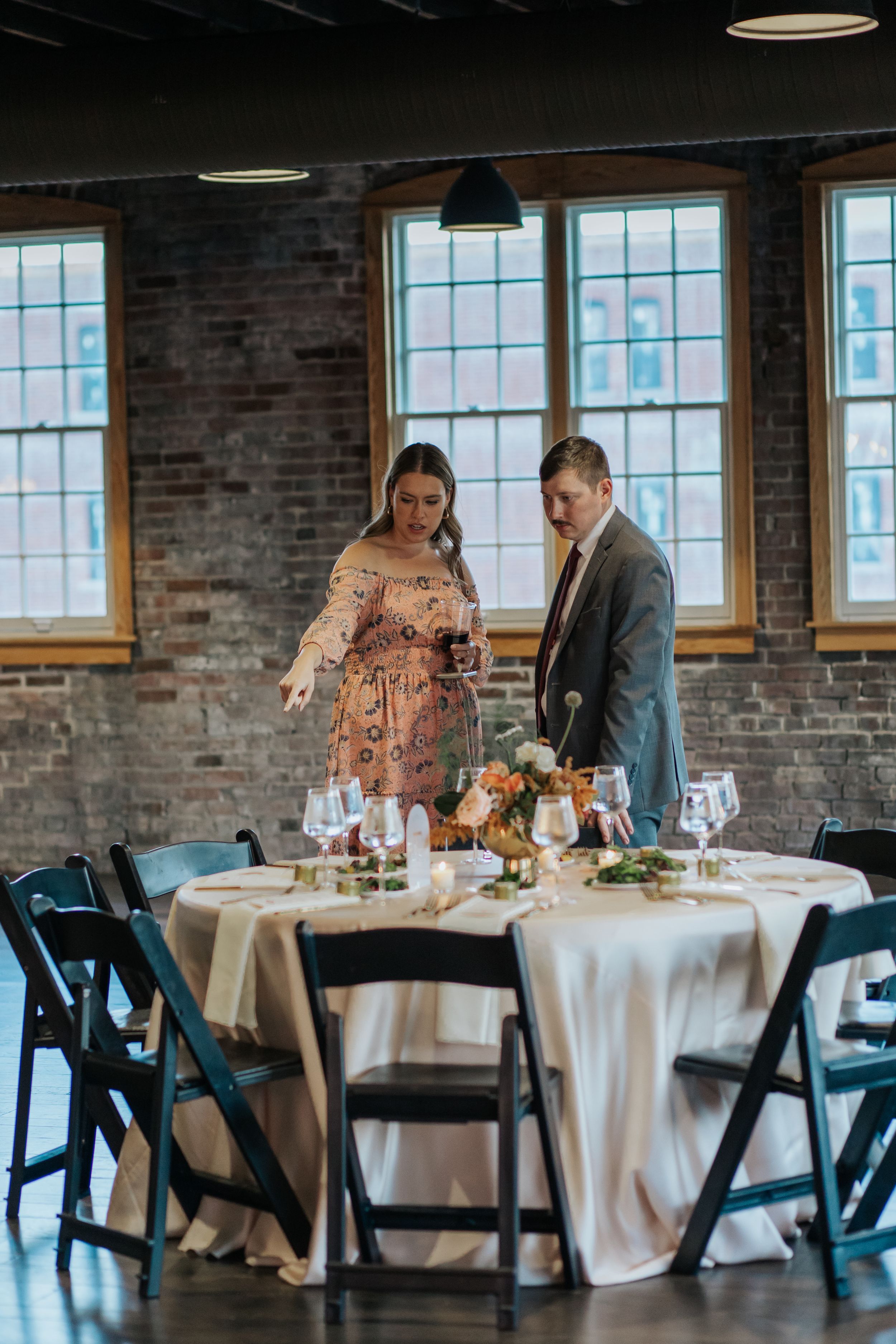 Wedding reception setup in industrial venue with exposed brick walls, white tablecloths and black chairs.