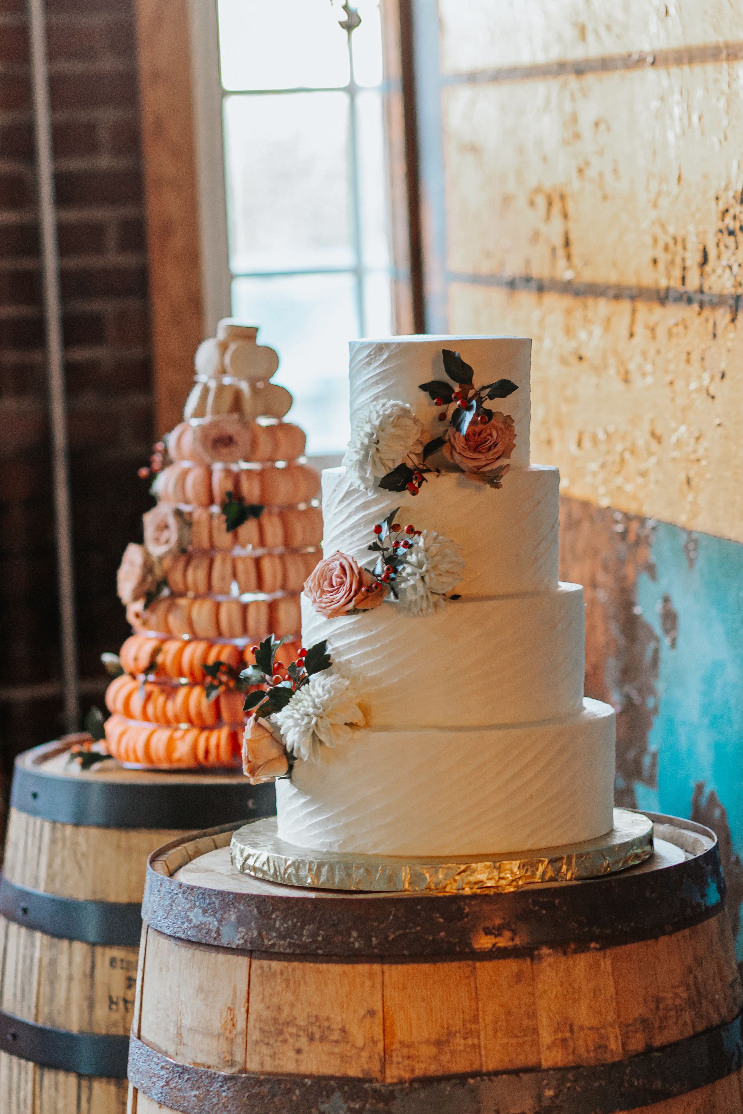 Three white wedding cakes with blush roses displayed on wooden barrel stands against a brick wall.