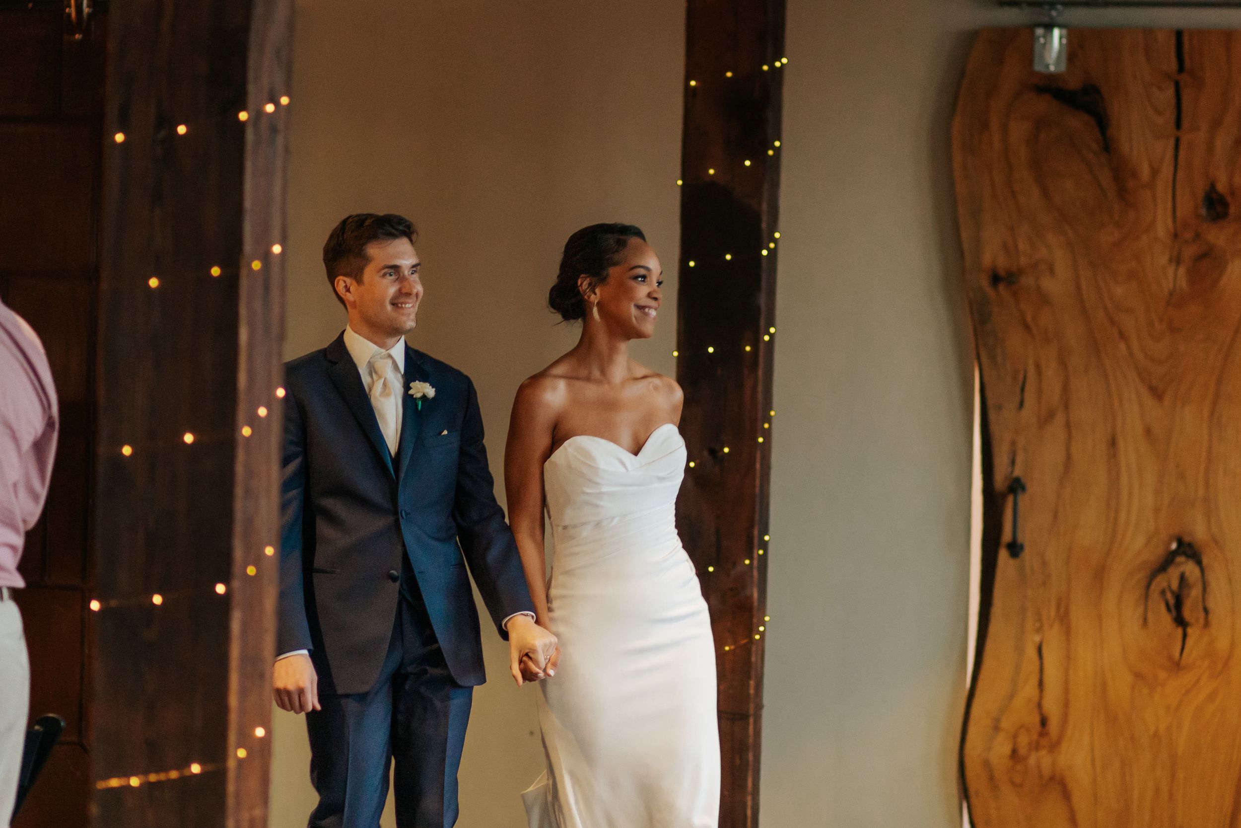 A bride and groom share their first dance at a rustic barn wedding reception with twinkling lights in the background.