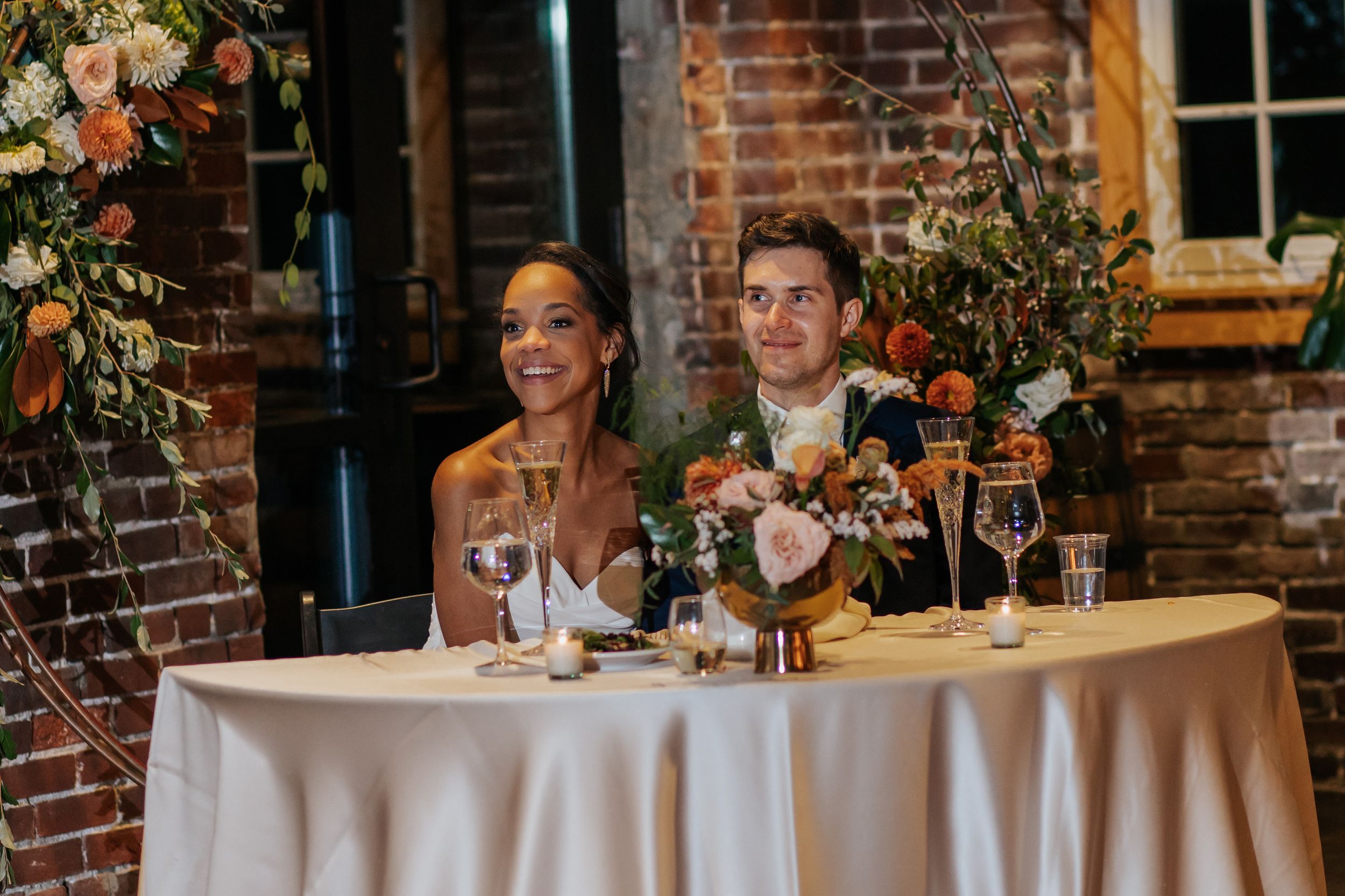 Guests seated at a long dining table in a rustic brick-walled venue with elegant place settings and wine glasses.