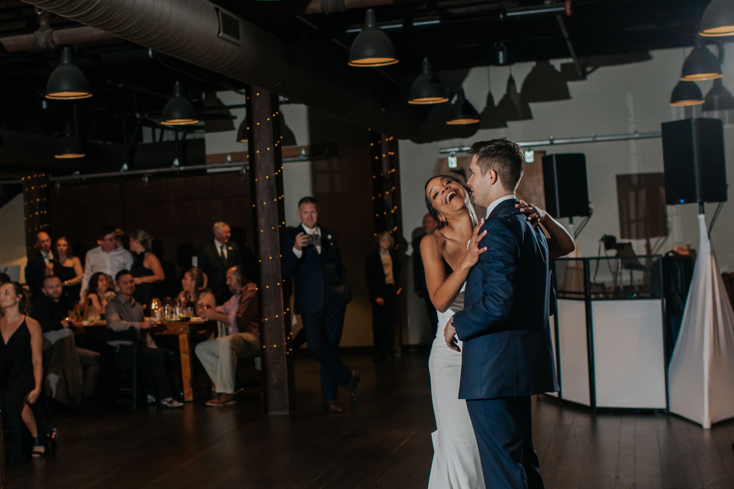 A couple shares their first dance at a dimly lit wedding reception venue while guests watch from their tables.