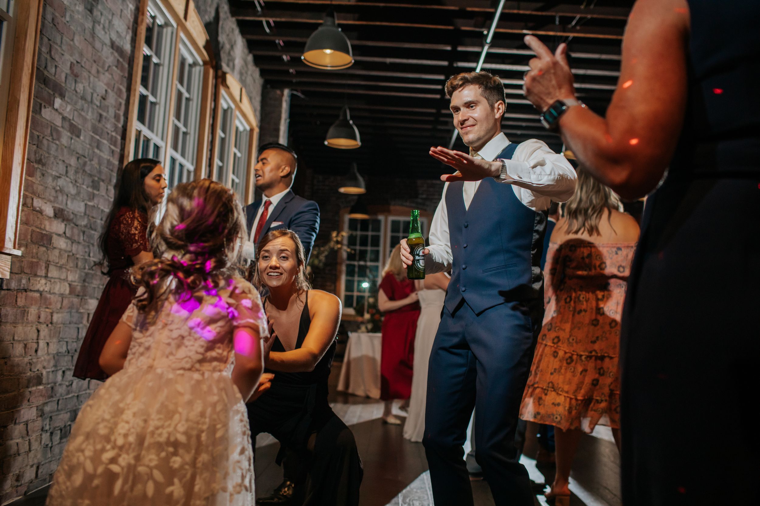 Wedding guests mingle and dance during reception in rustic brick-walled venue with pendant lighting.