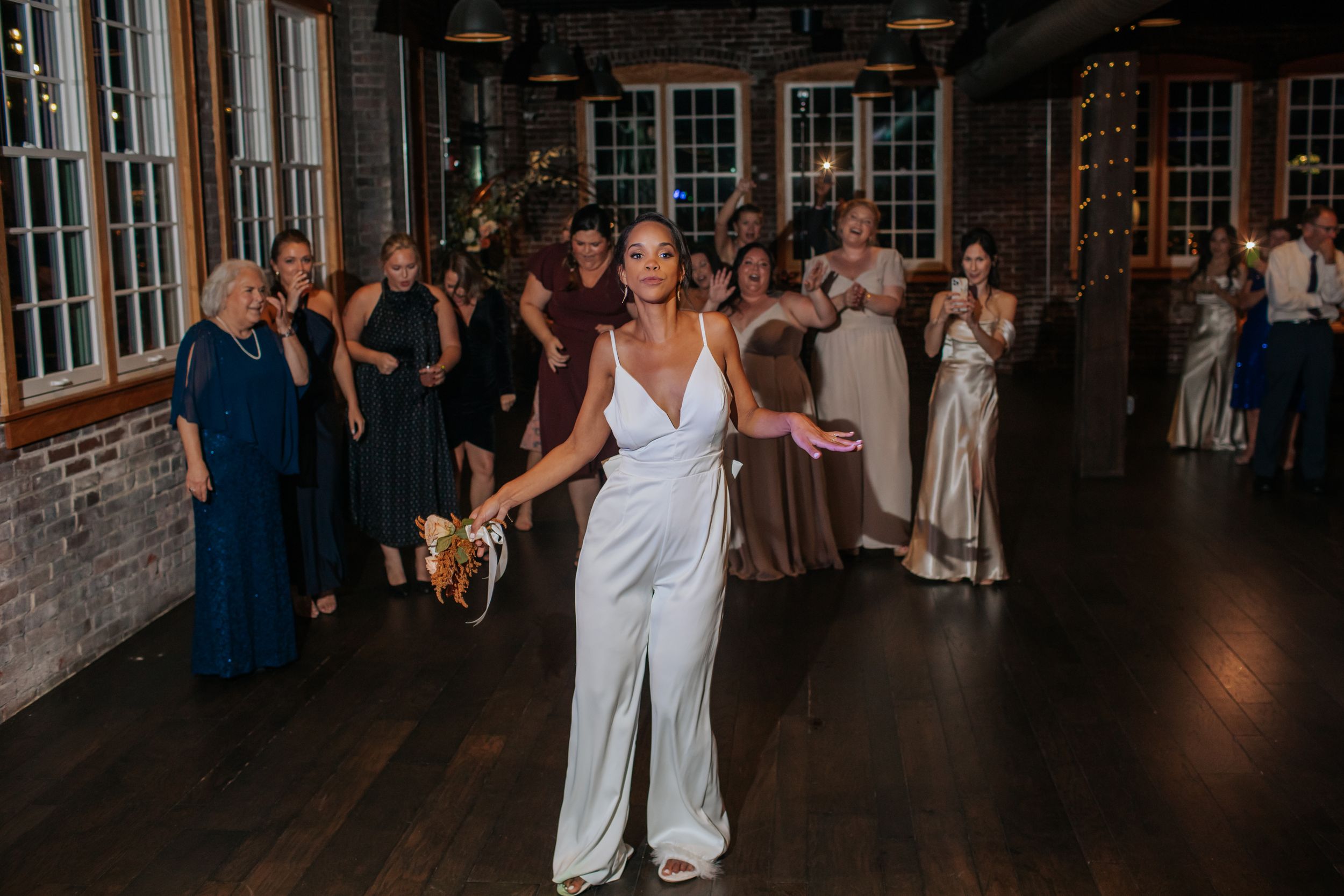 A person in a white jumpsuit dances joyfully at a wedding reception while guests watch in the background.