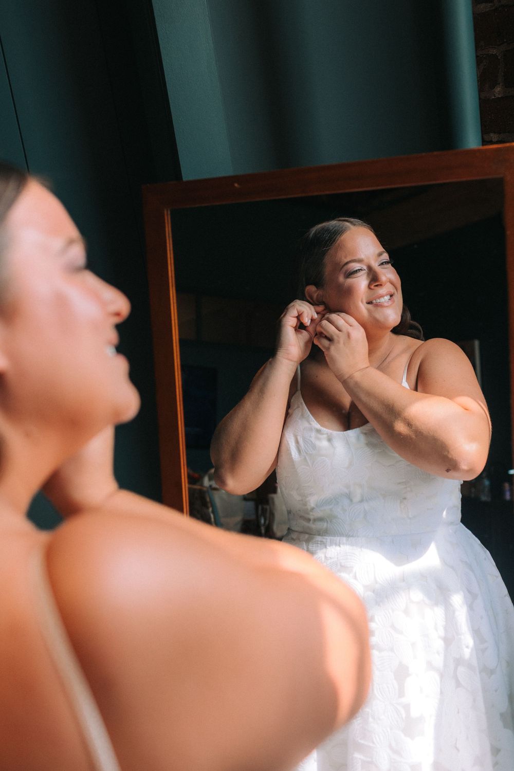A joyful person in a white wedding dress puts on earrings while looking in a mirror with a bright smile.