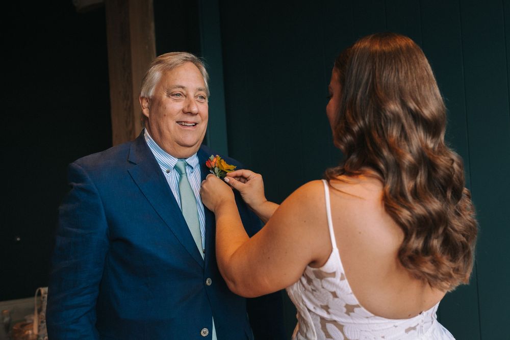 Someone helps adjust a boutonniere on a person wearing a blue suit jacket during a formal event.