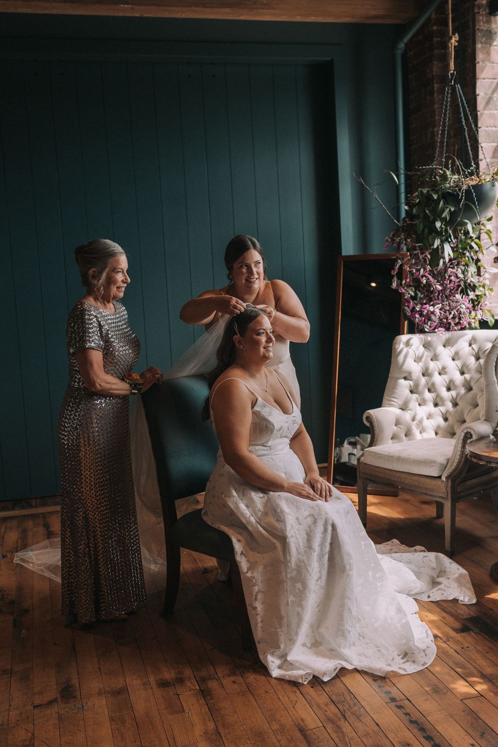 A bride in a white lace gown gets ready in a rustic room with wooden floors and teal walls.