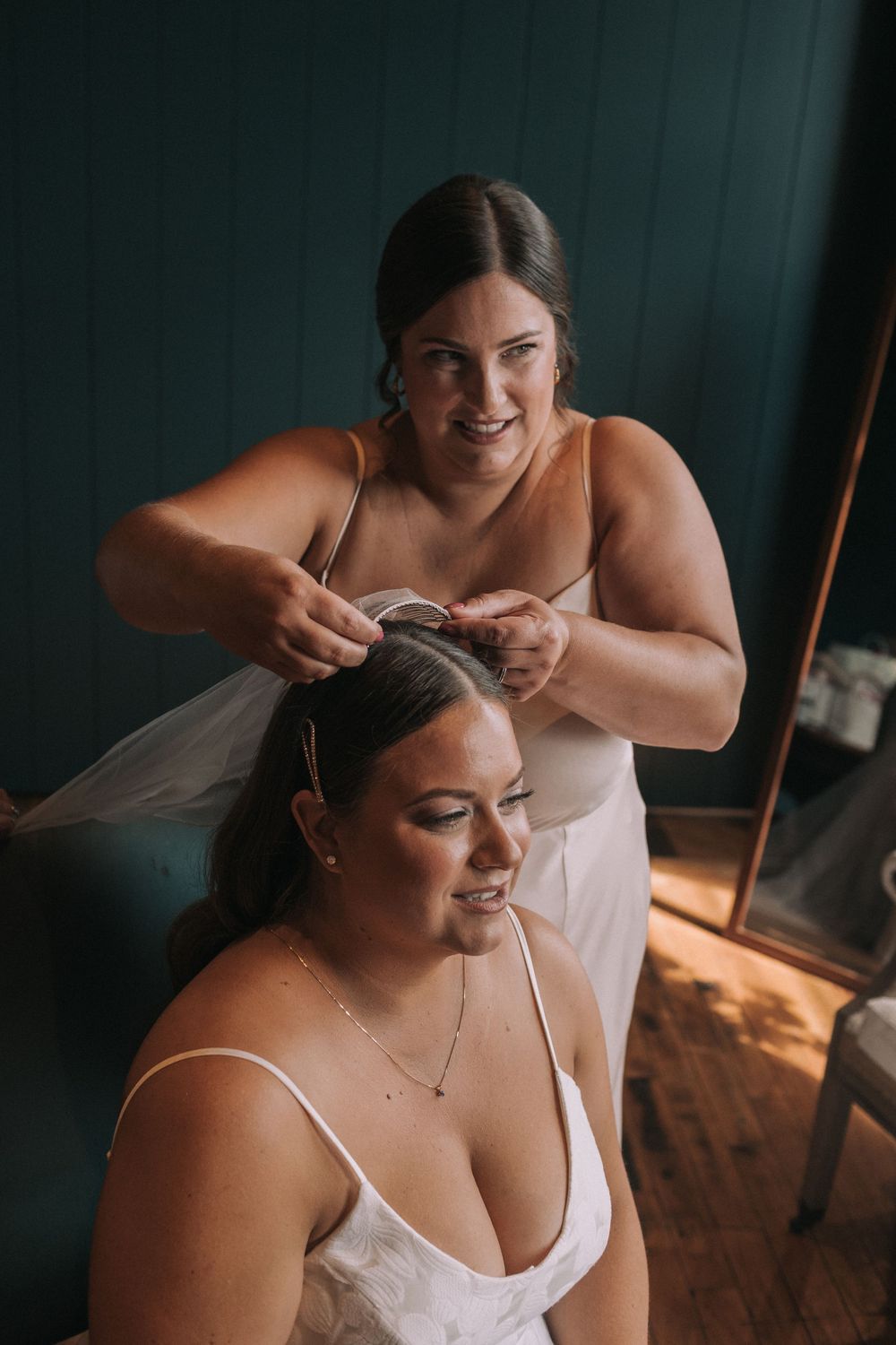 Two people in white dresses share an intimate moment during wedding preparations as one helps style the other's hair.