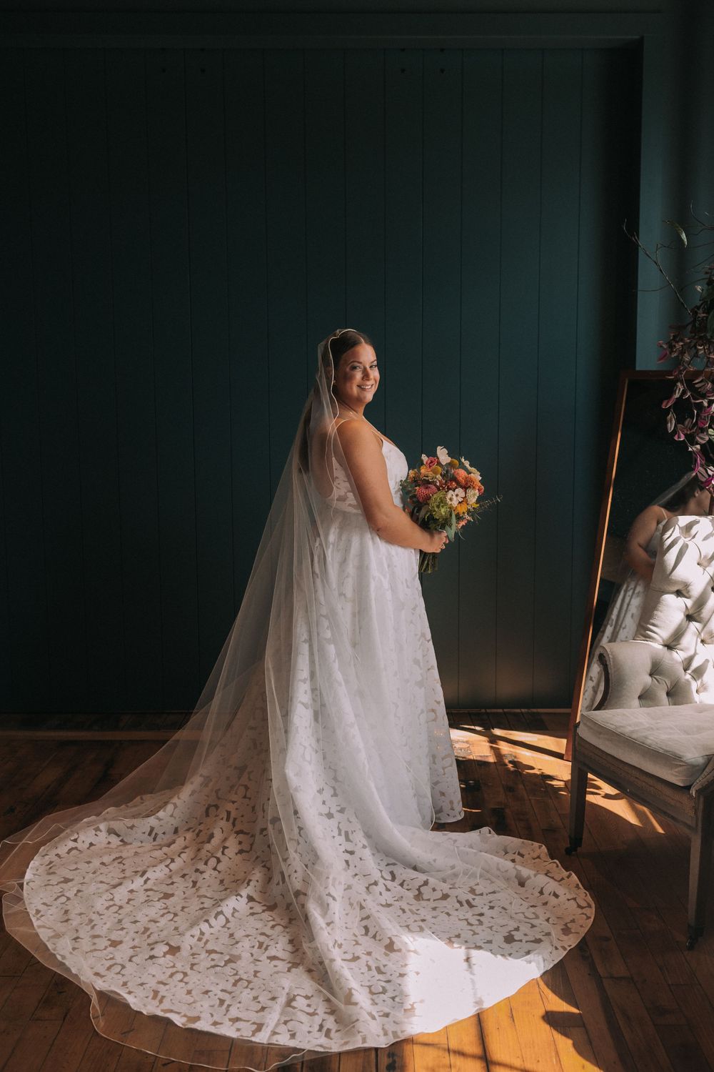 A bride in a flowing white lace wedding gown with a dramatic cathedral train sits gracefully in natural window light.