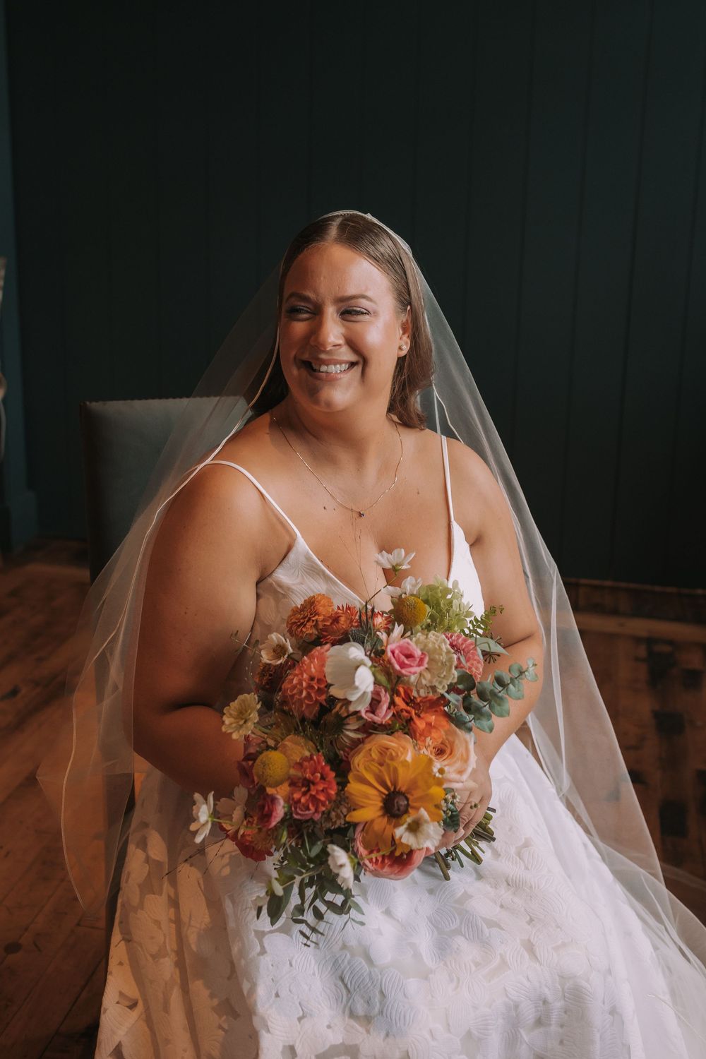 A bride in a white wedding dress holds a vibrant autumn-colored bouquet featuring sunflowers and orange blooms.