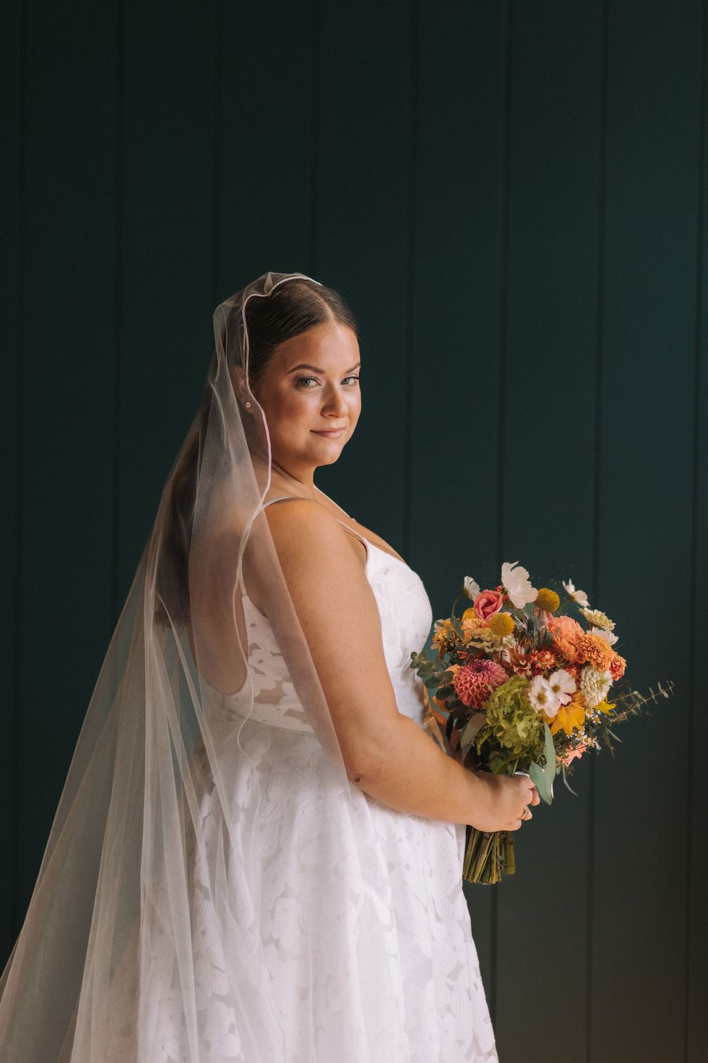 A bride in a white wedding dress holds a vibrant autumn bouquet against a dark green backdrop.