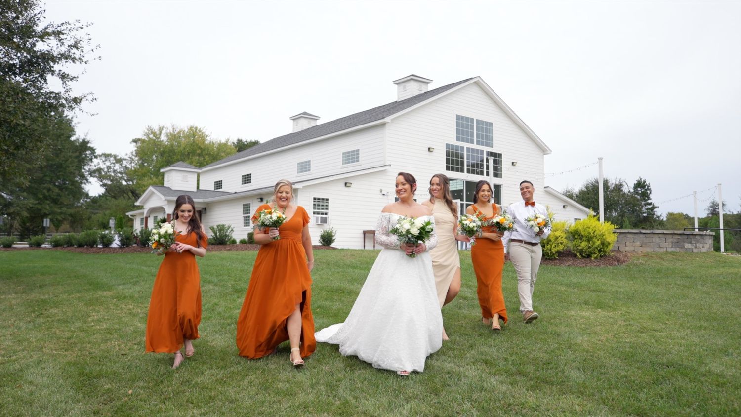 A wedding party in orange attire poses playfully on a grassy lawn in front of a white building.