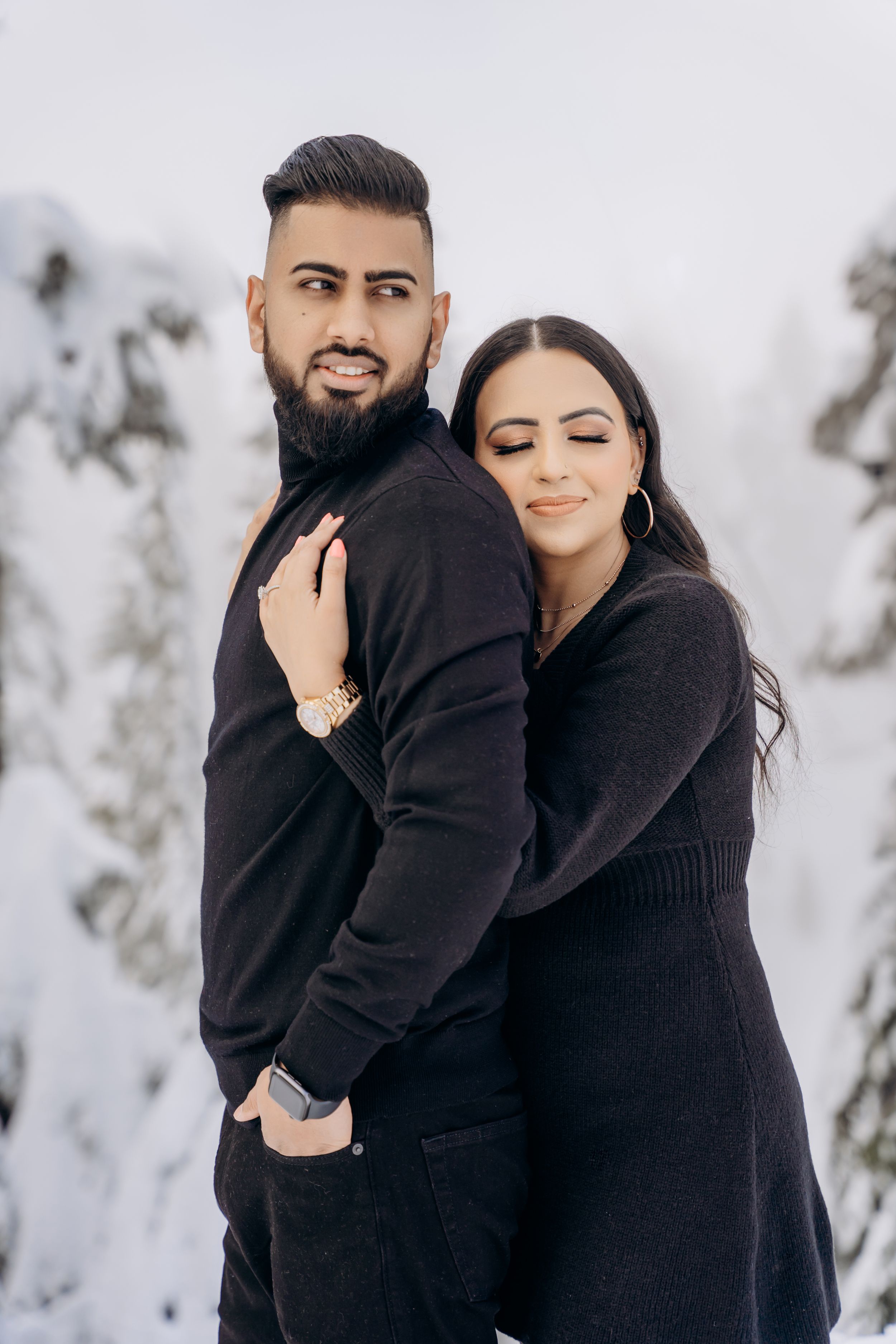 A couple wearing matching black sweaters embrace in a romantic winter photoshoot with snowy scenery.