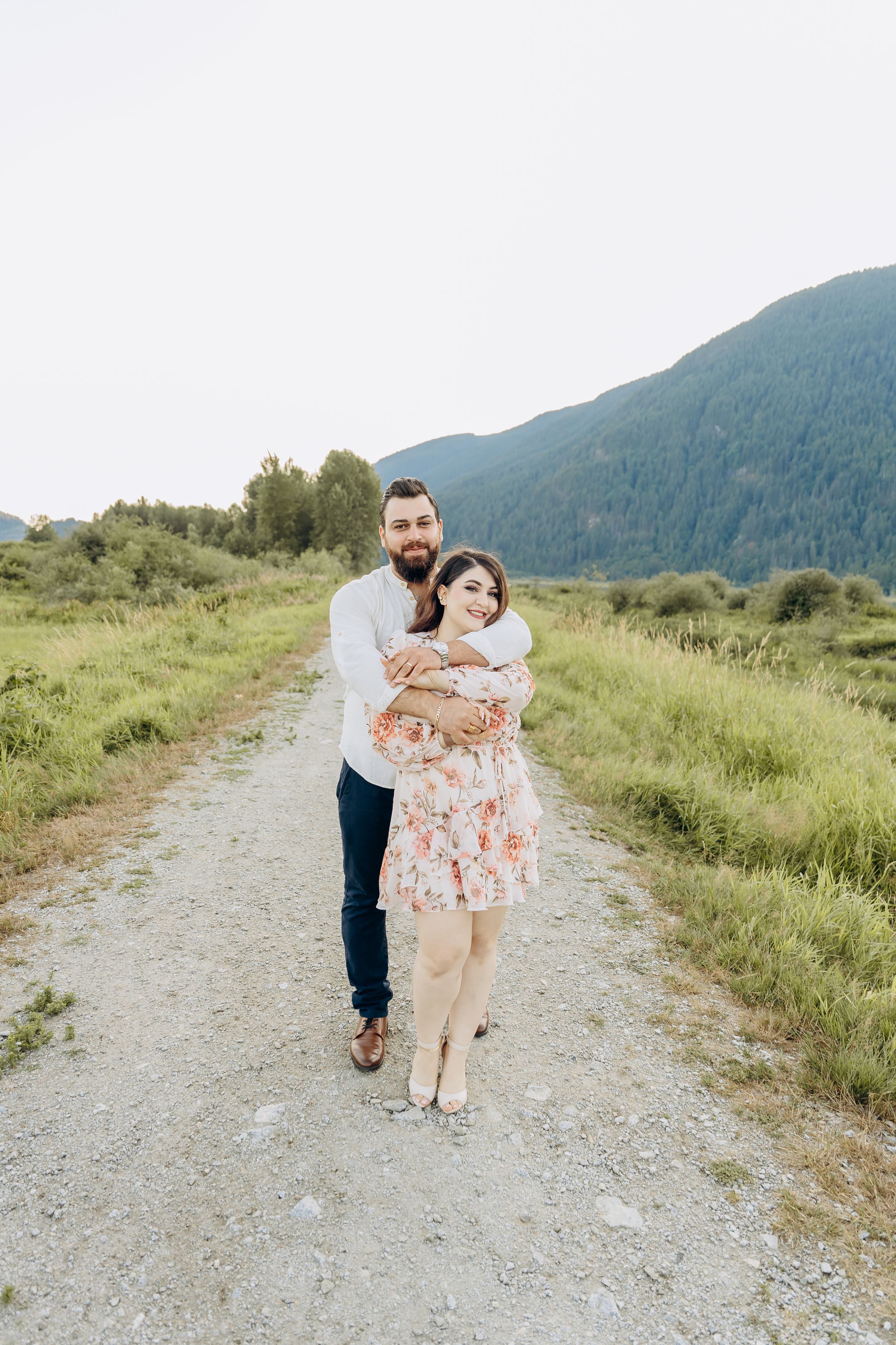 A couple stands together on a gravel path with green mountains and meadows in the background.
