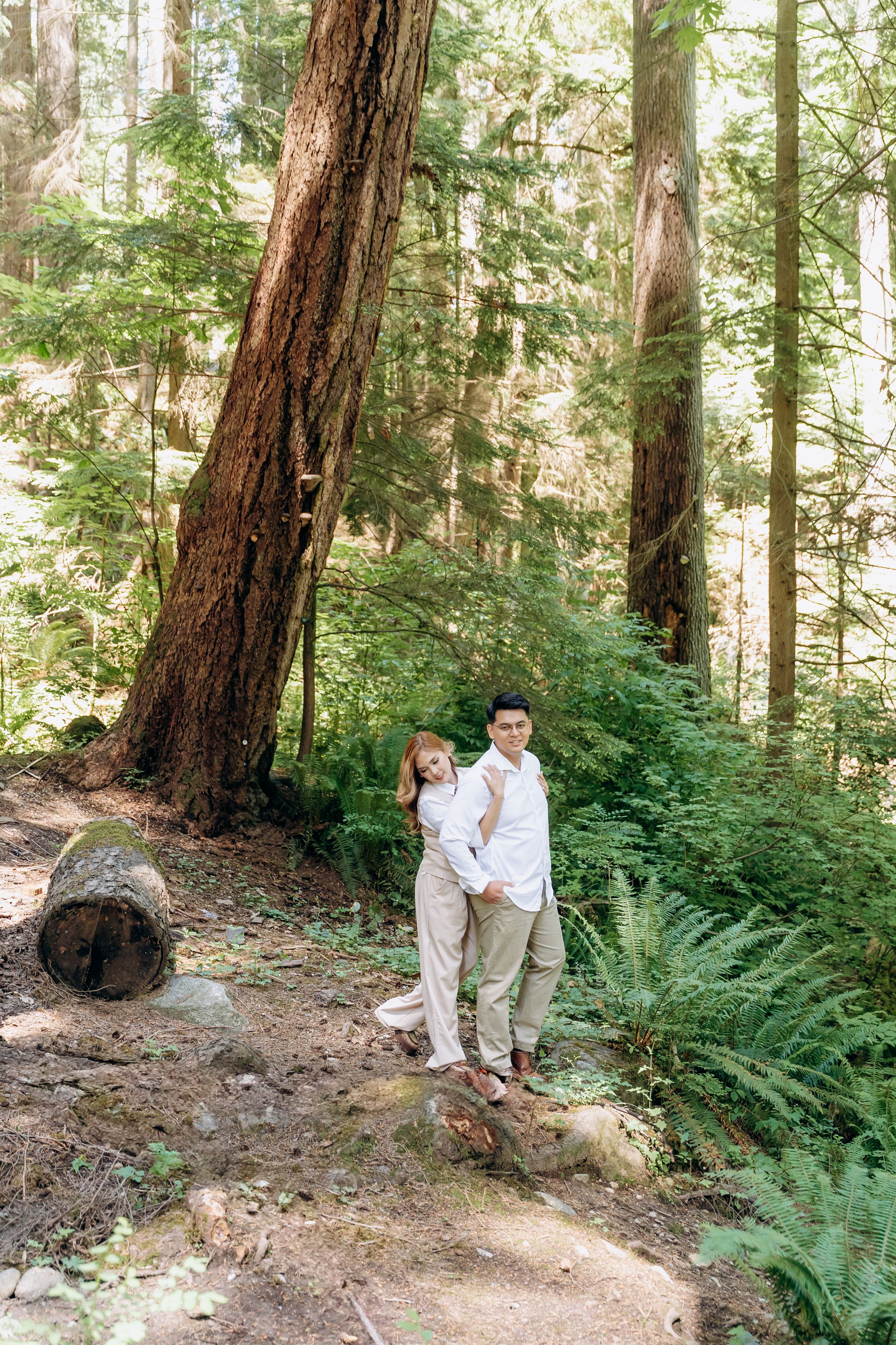 A couple shares an intimate moment among tall redwood trees and ferns in a peaceful forest setting.