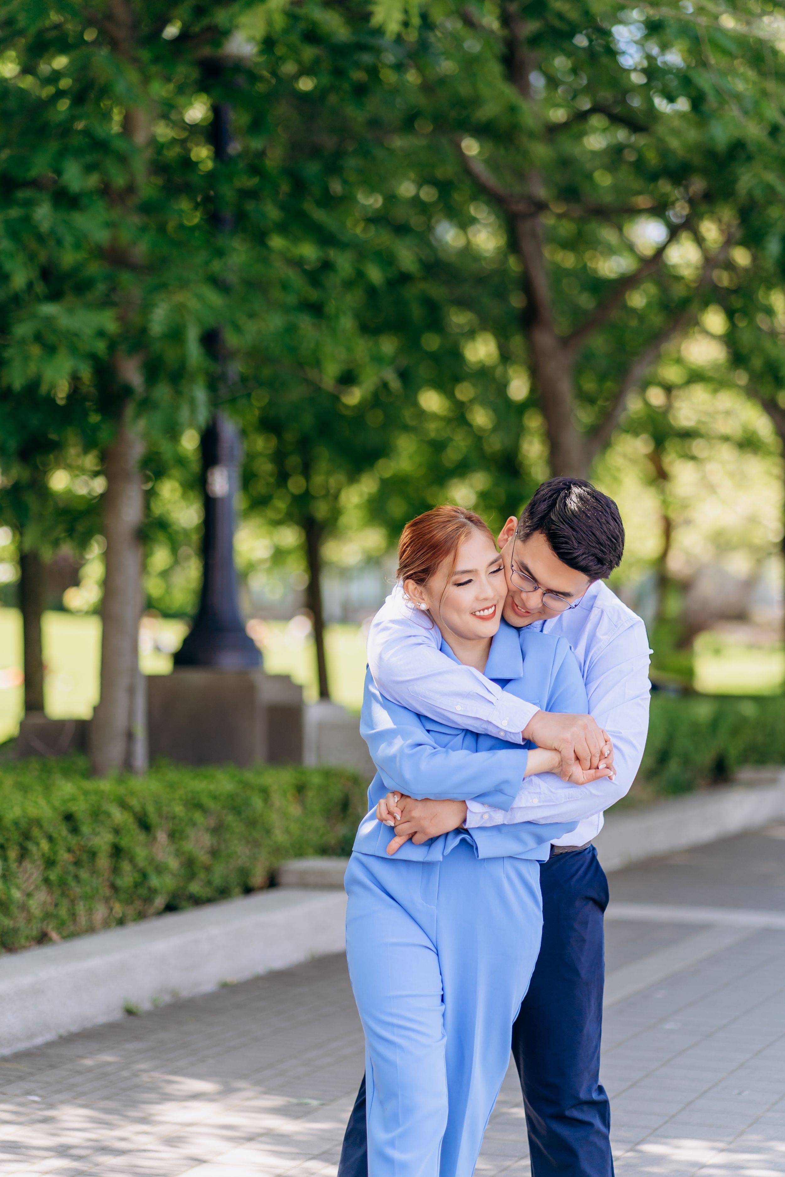 A couple shares a tender moment while embracing in a sunny park with trees in the background.
