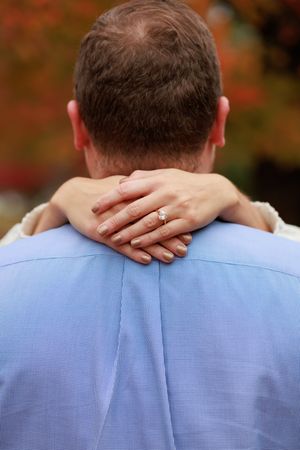 A woman's hand rests gently on the back of someone wearing a light blue dress shirt against an autumn background.