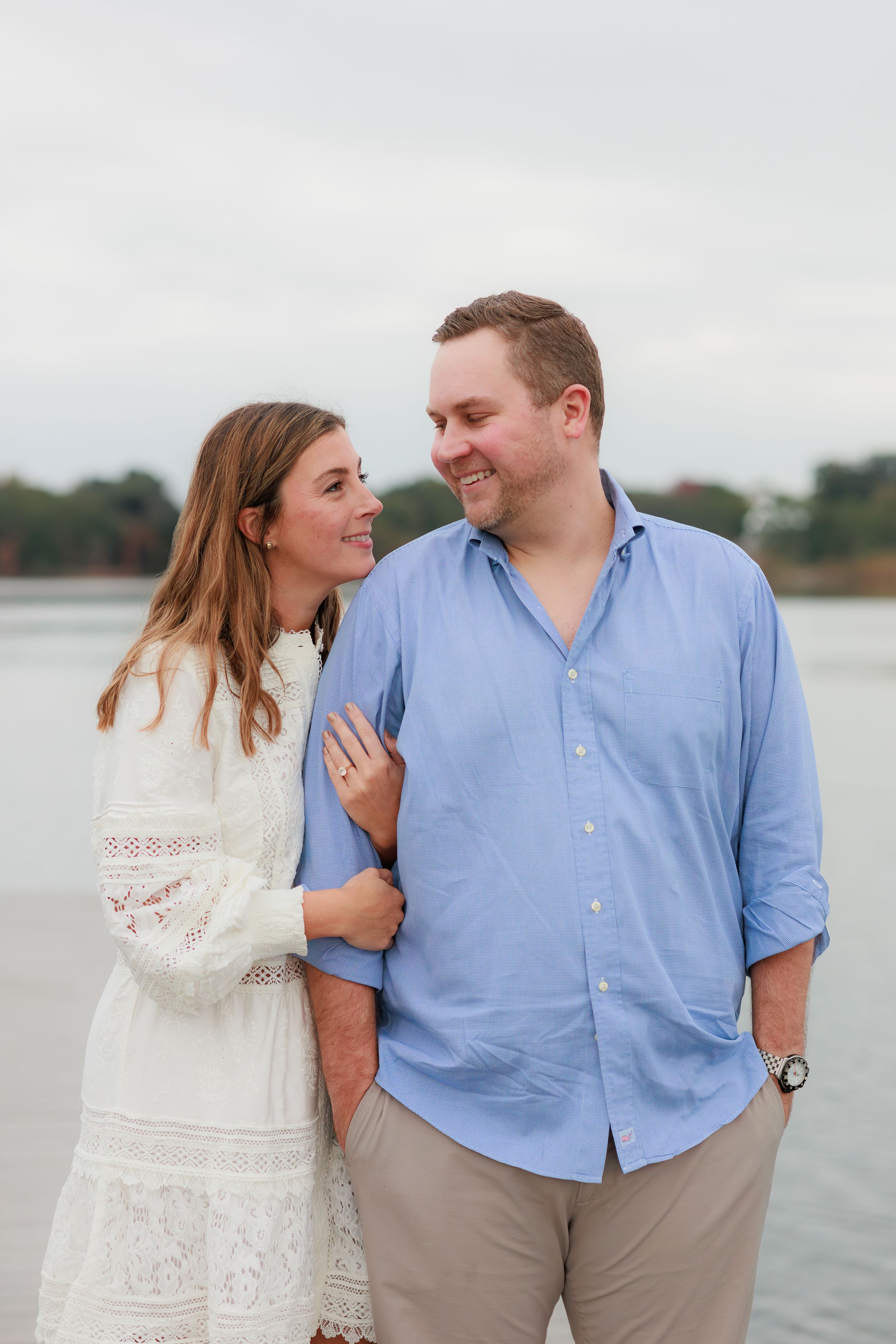 A couple shares a tender moment by the water, with her in a white lace dress and him in a light blue button-down shirt.