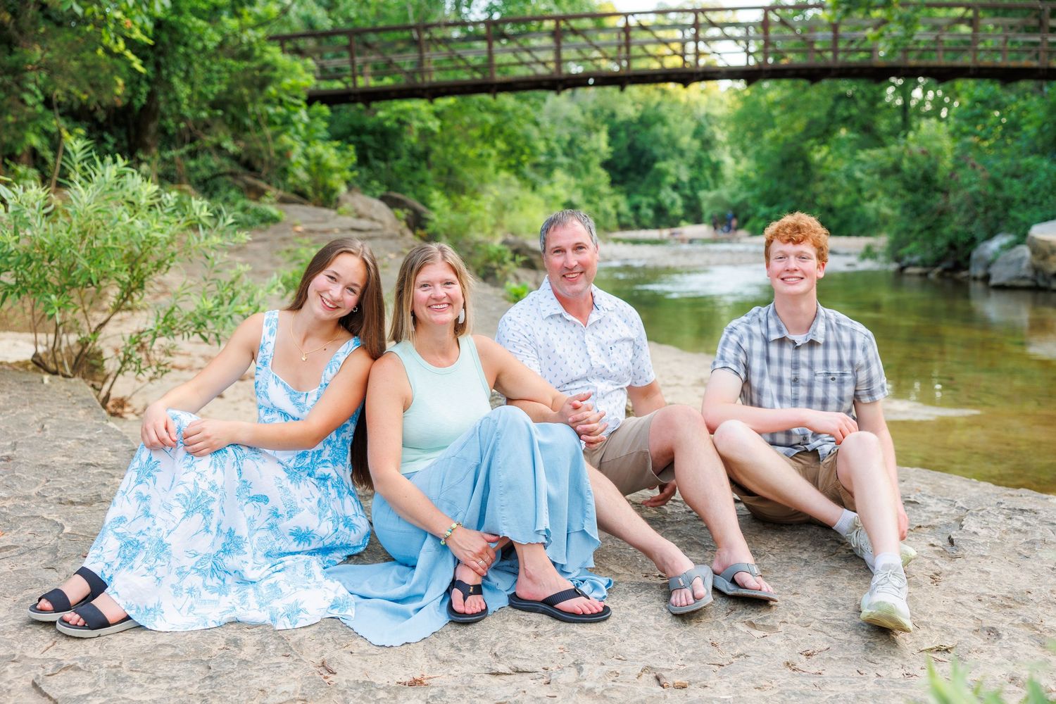 Family in coordinated blue and white outfits relaxing by a creek during an outdoor photo session.