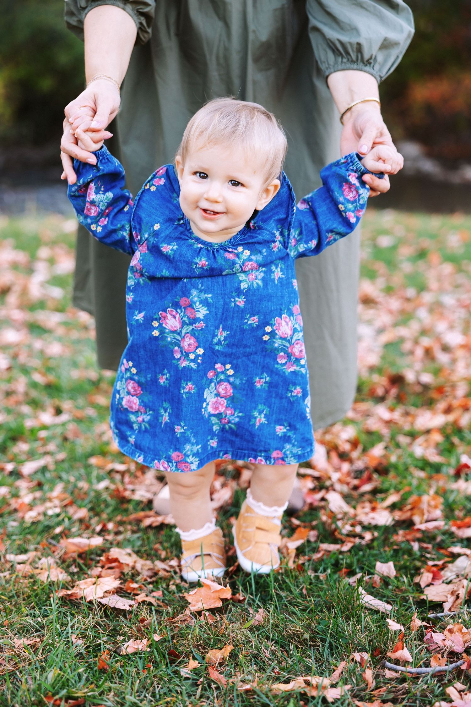 Small child wearing bright blue floral dress stands in autumn leaves while holding adult's hands.