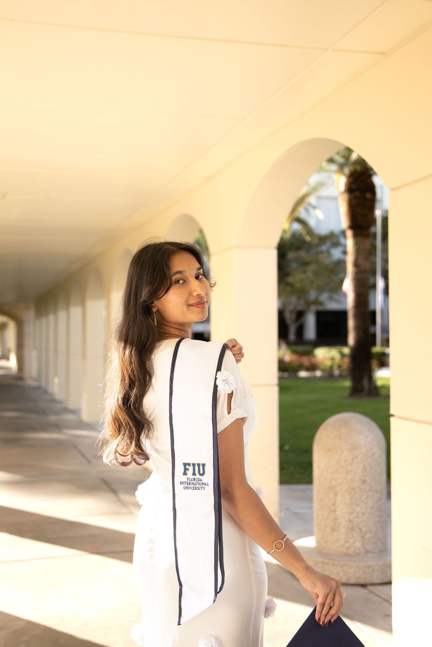 Student wearing white FIU shirt poses in outdoor campus colonnade with sunlit architectural columns and arches.