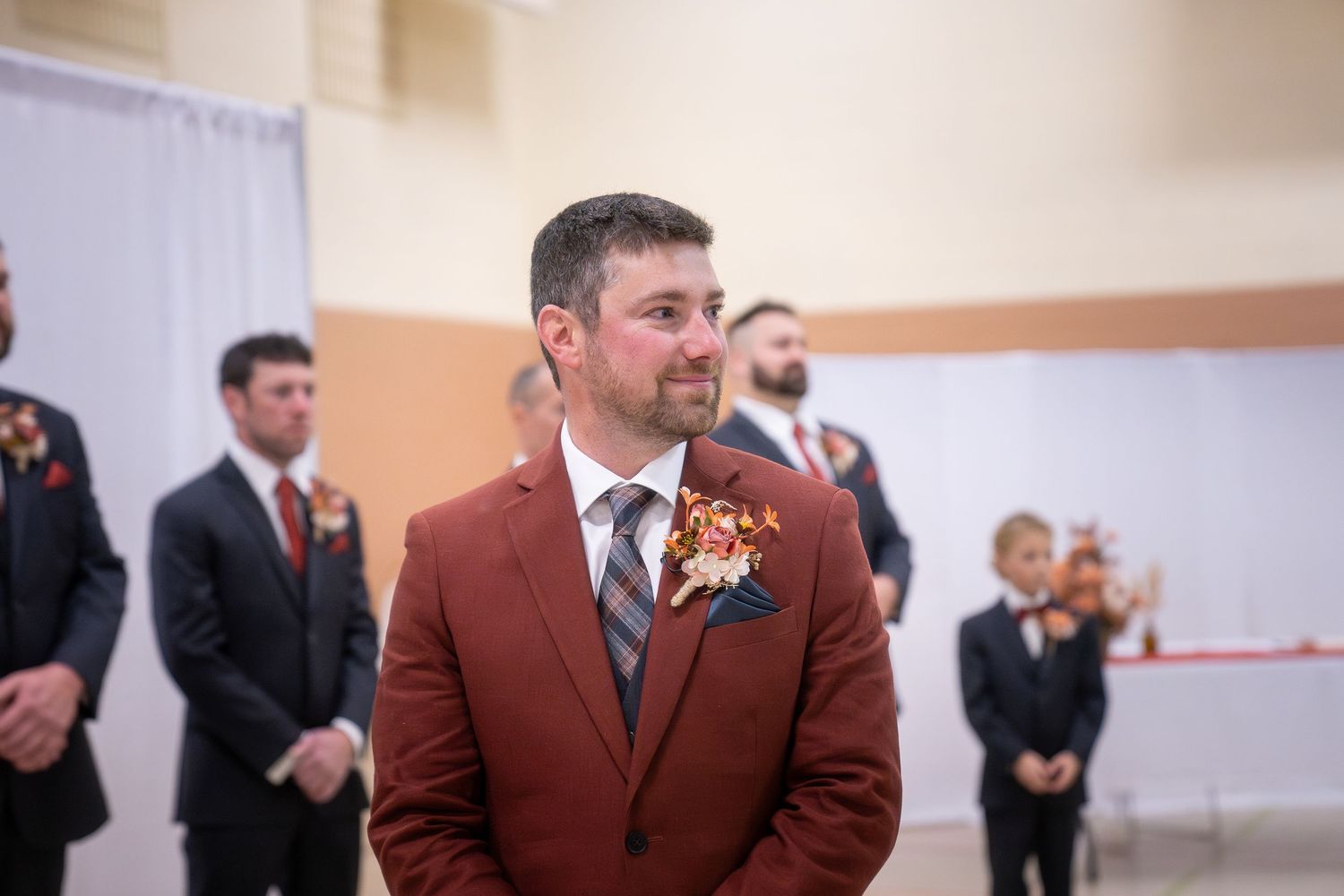 A groom in a rust-colored suit with boutonniere stands during wedding ceremony while groomsmen wait in background.