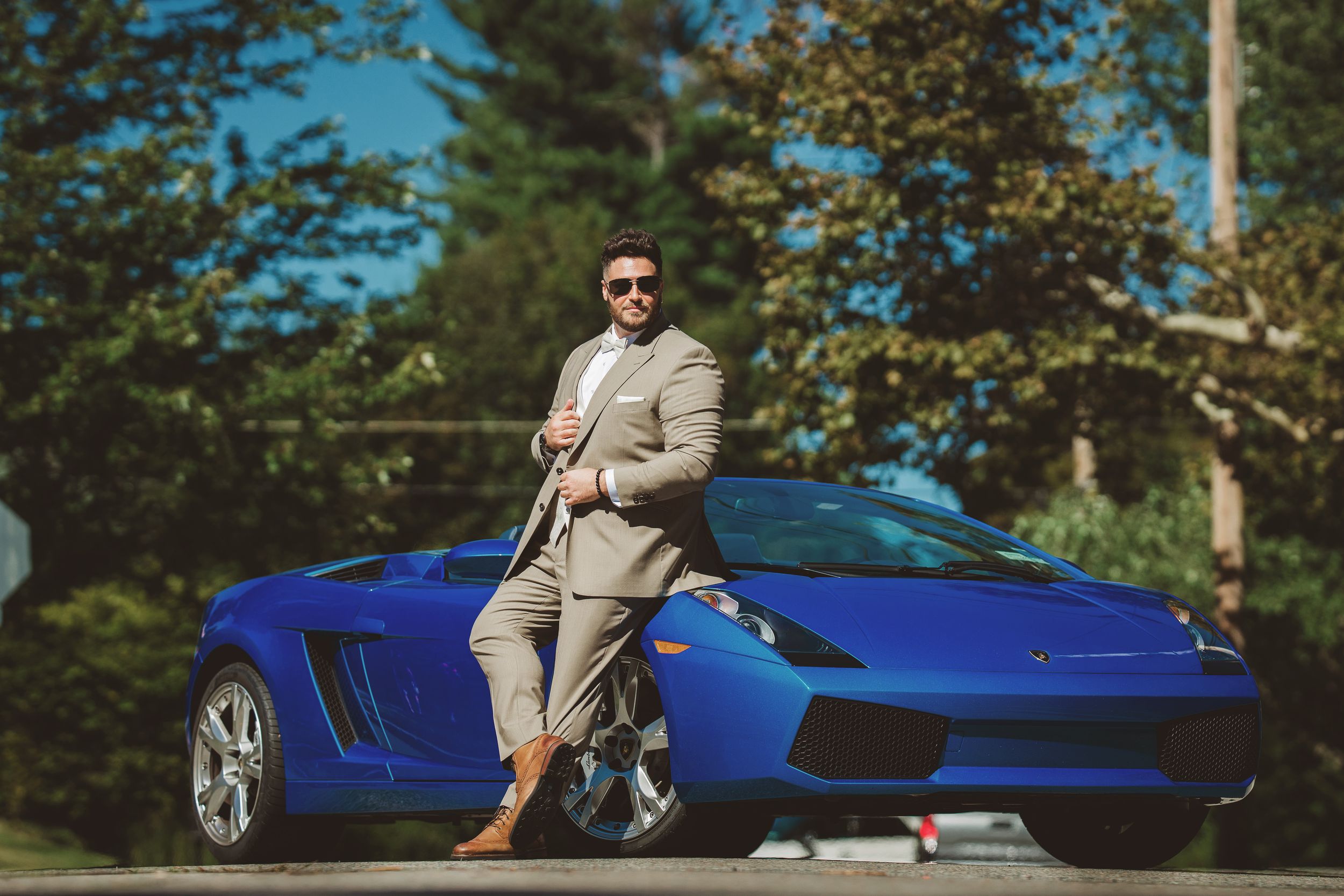 A bright blue sports car Lamborghini is parked in a sunny outdoor setting with trees and greenery in the background for groom prep.