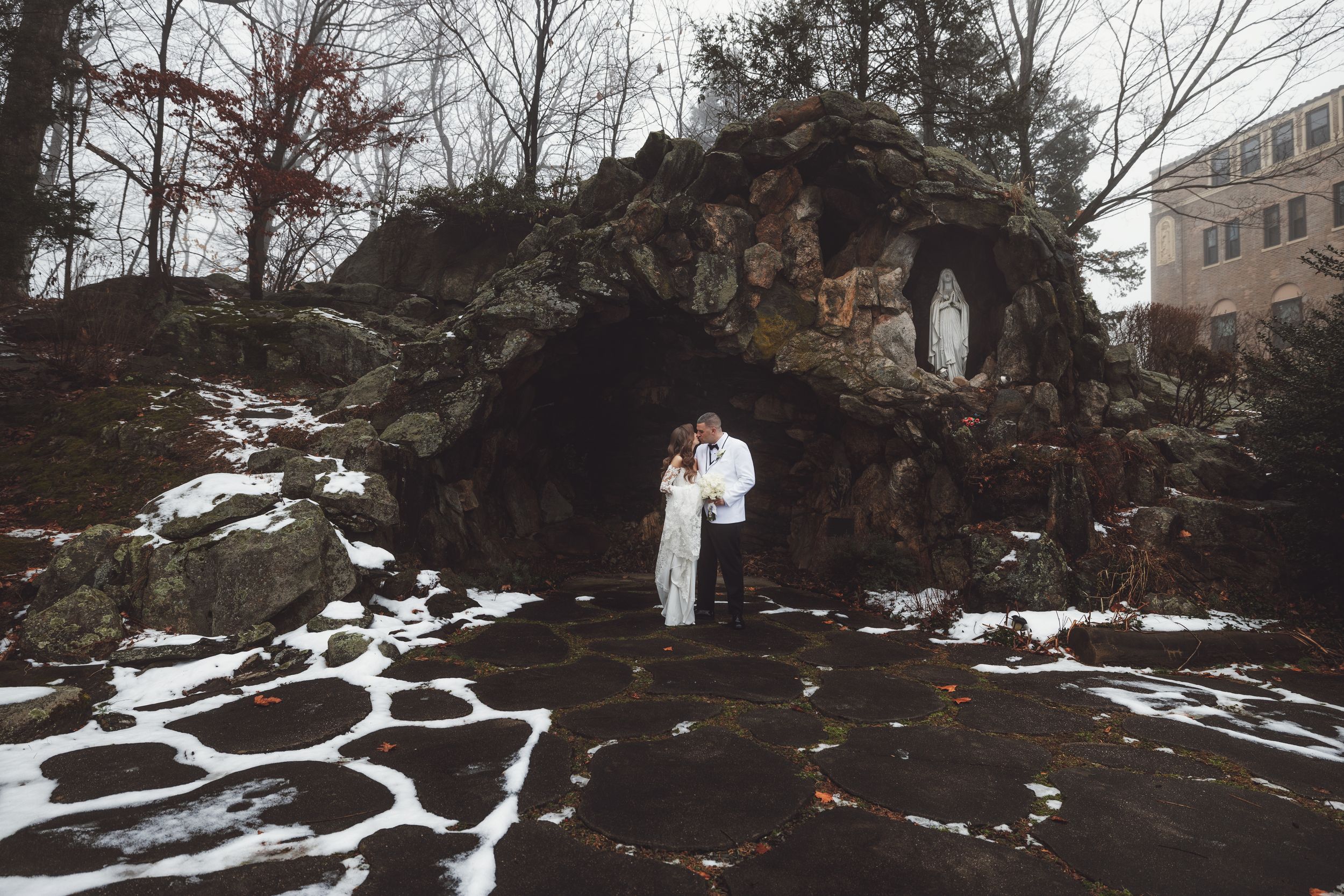 A winter wedding photo with a couple standing in front of a snow-covered stone grotto.