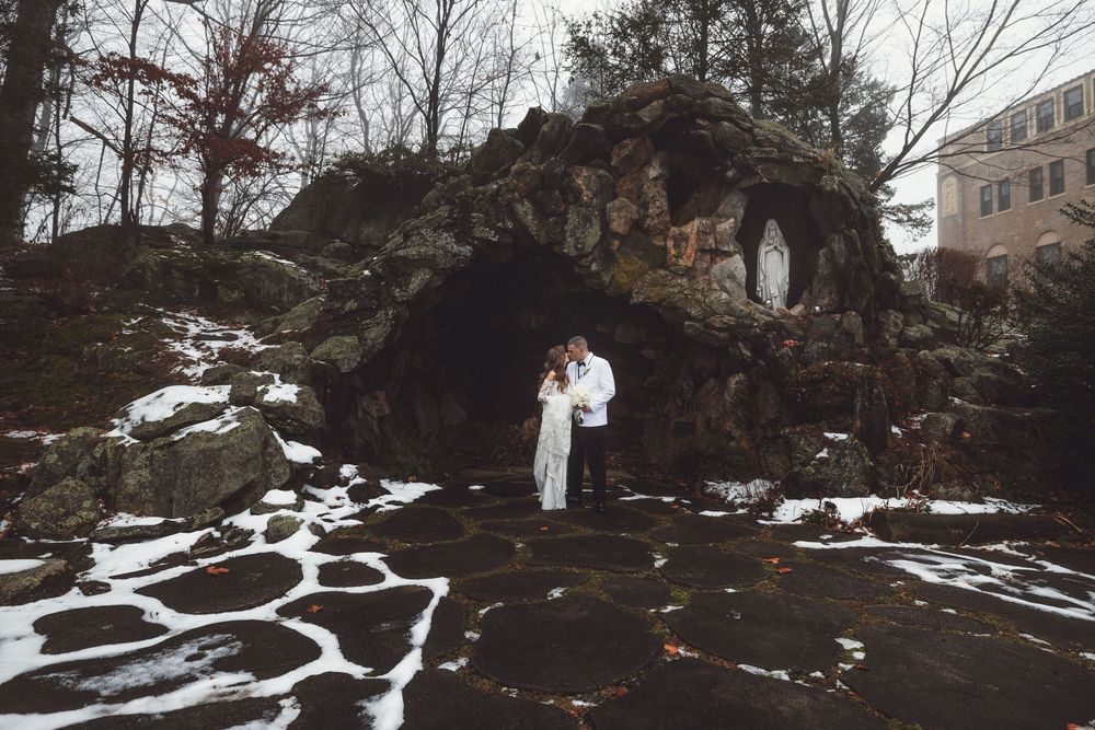 A winter wedding photo with a couple standing in front of a snow-covered stone grotto.