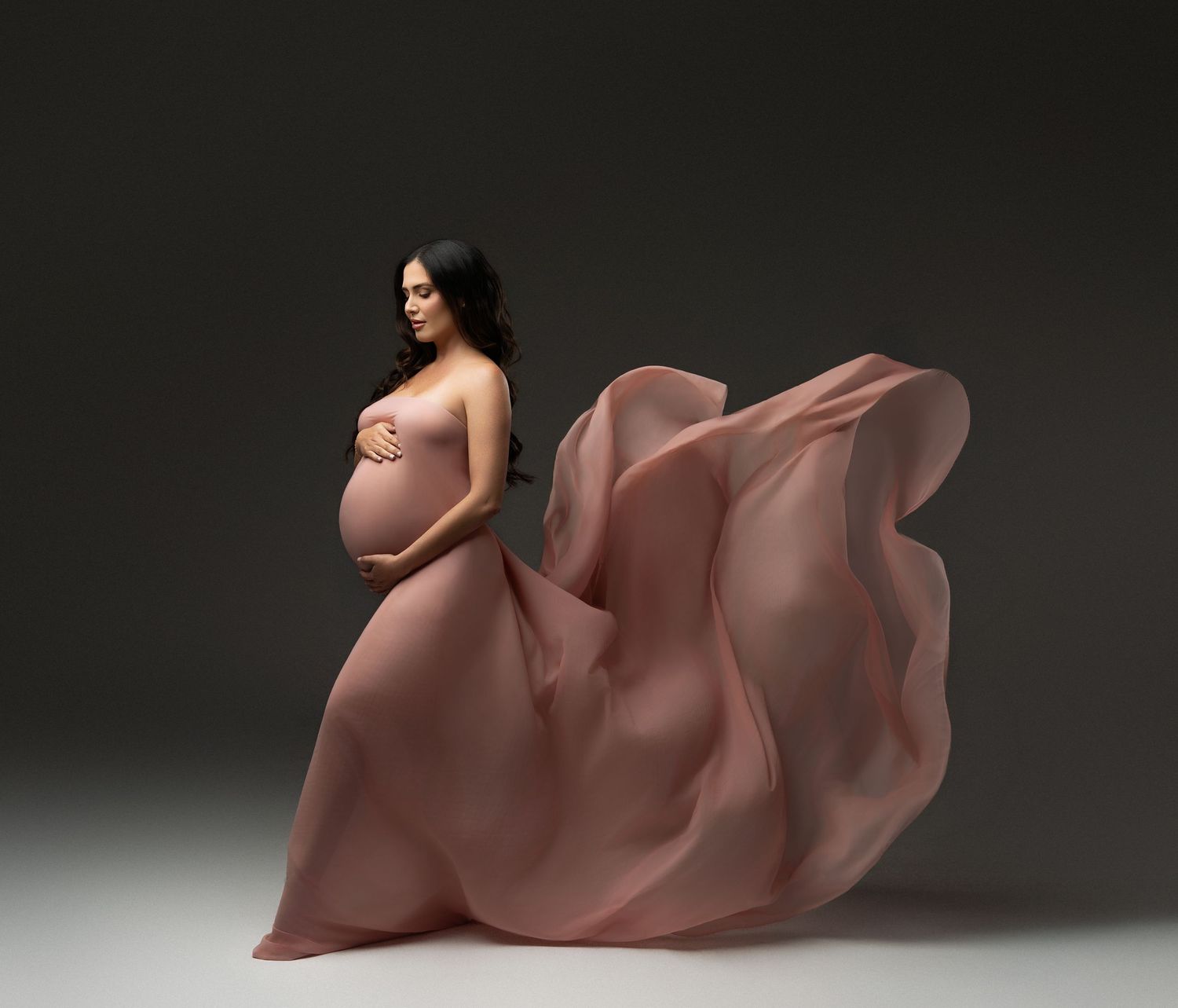 A flowing red maternity gown billows dramatically against a dark background in an elegant pregnancy portrait.