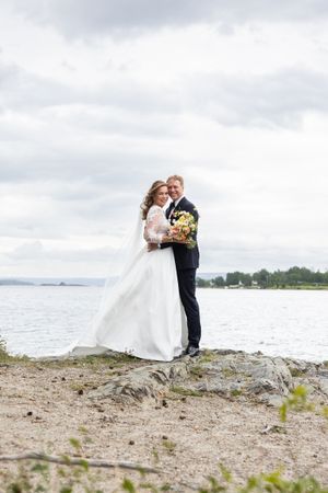 Brudeparet står på en liten topp, bak dem er det vann og dramatiske skyer.