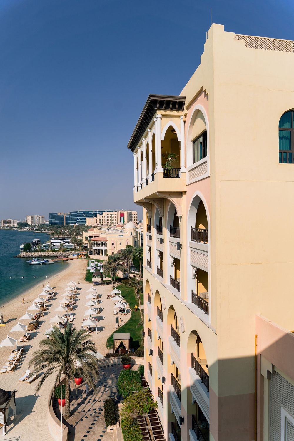 Luxury beachfront hotel with Mediterranean architecture overlooks sandy beach with umbrellas along Qatar's coastline.