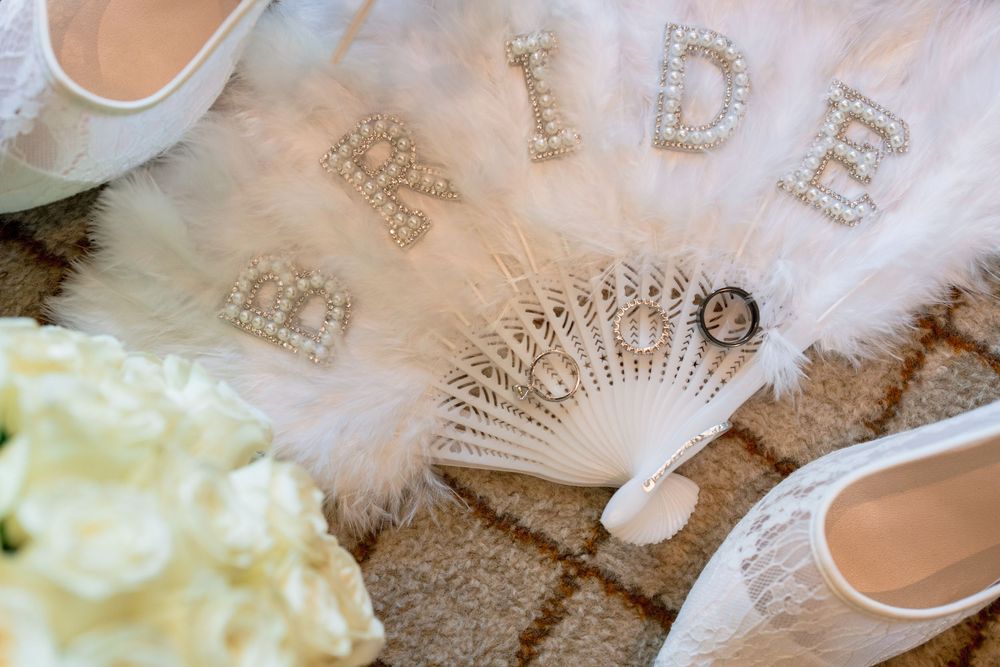 Wedding accessories including a white feather fan, sparkly 'BRIDE' text, and white shoes arranged on a beige carpet.
