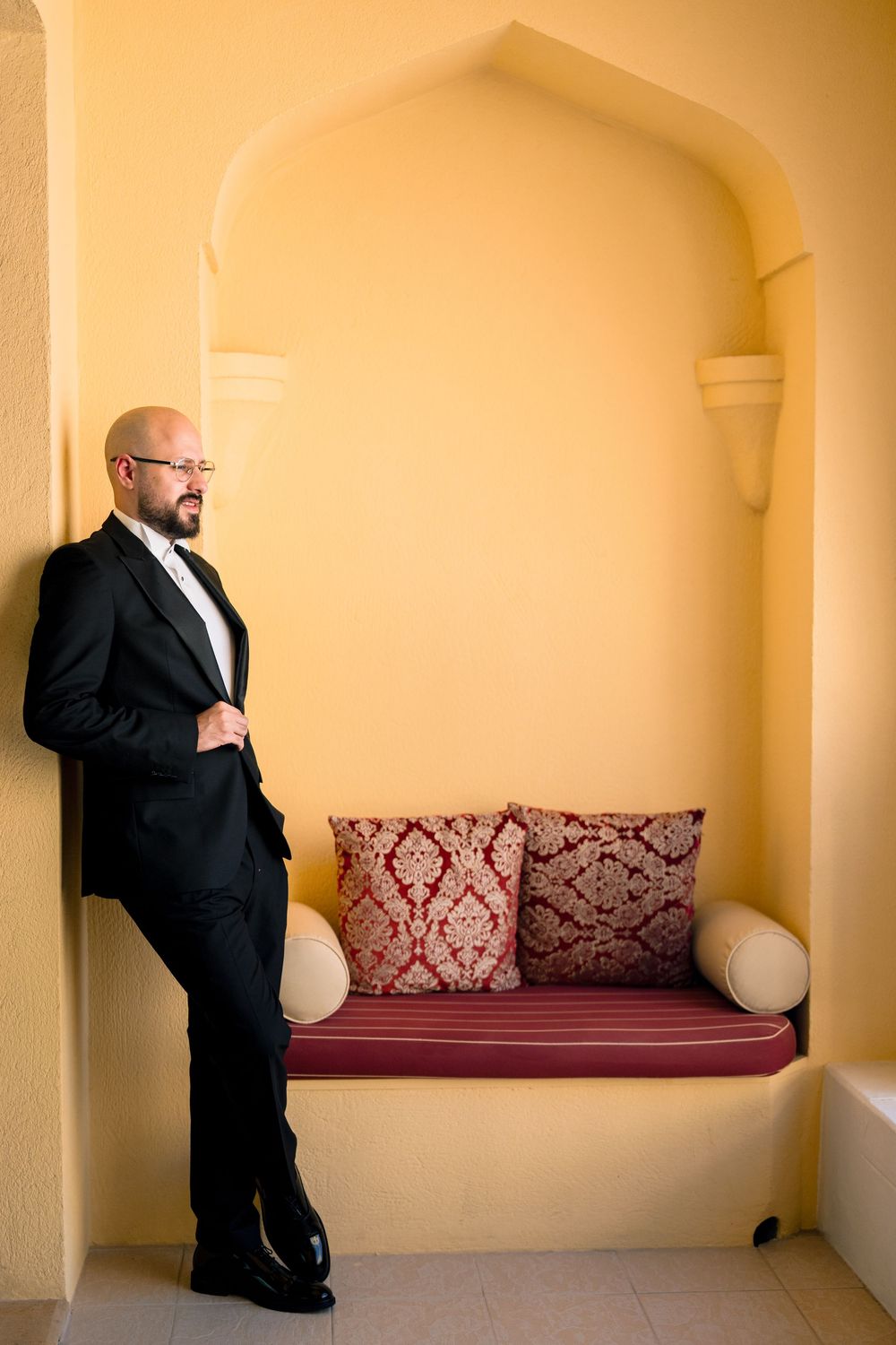 A person in a black suit stands in front of a built-in bench with burgundy cushions in an arched alcove.
