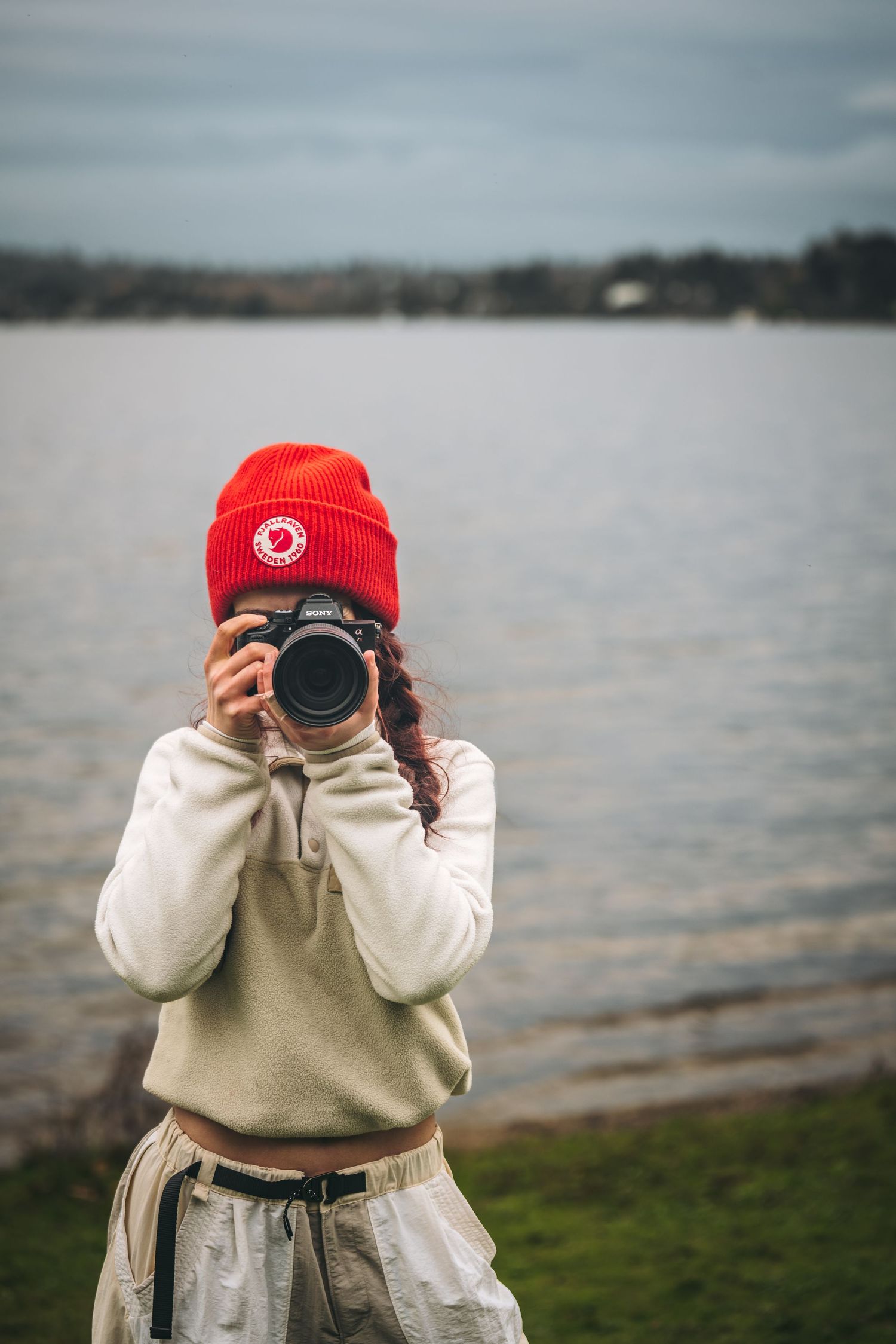 Woman in red beanie hat photographing with Sony camera by a lake on a cloudy day.