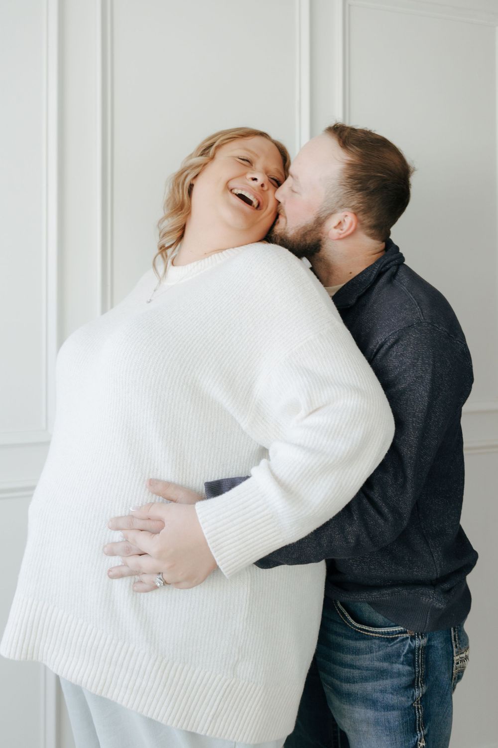 A couple in casual attire sharing a joyful embrace and laughing together against a white wall.