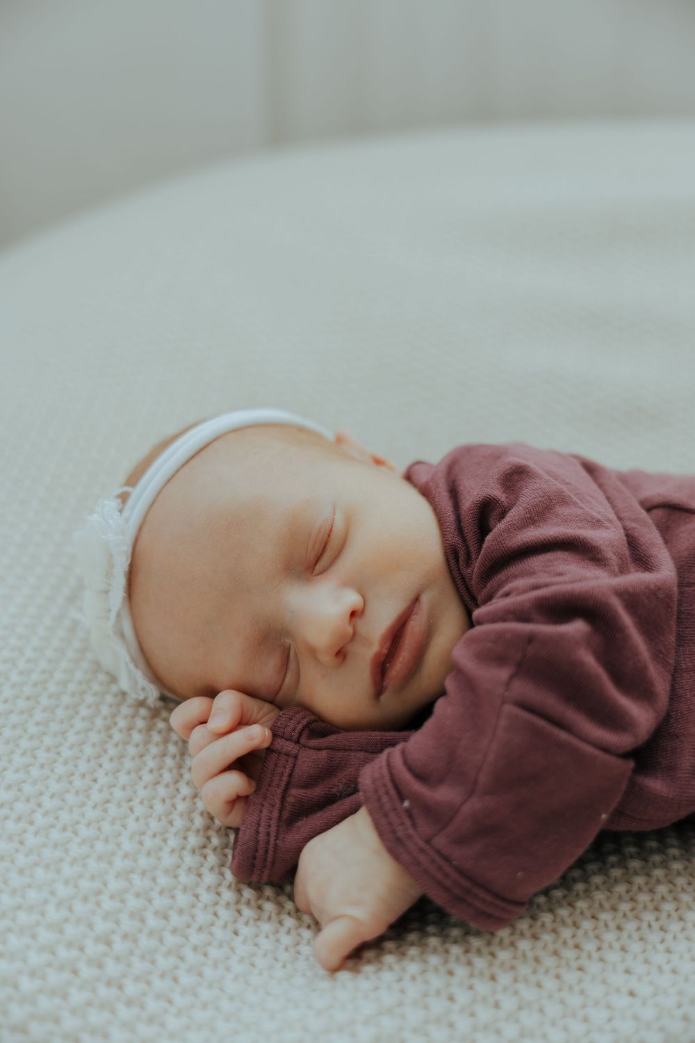 A sleeping newborn baby wearing a brown onesie rests peacefully on a textured white blanket.