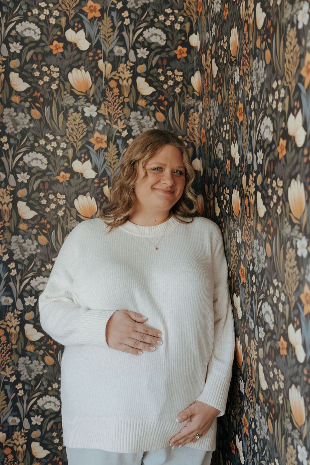 A series of pregnancy photos showing a person in a white sweater against a dark floral wallpaper background.