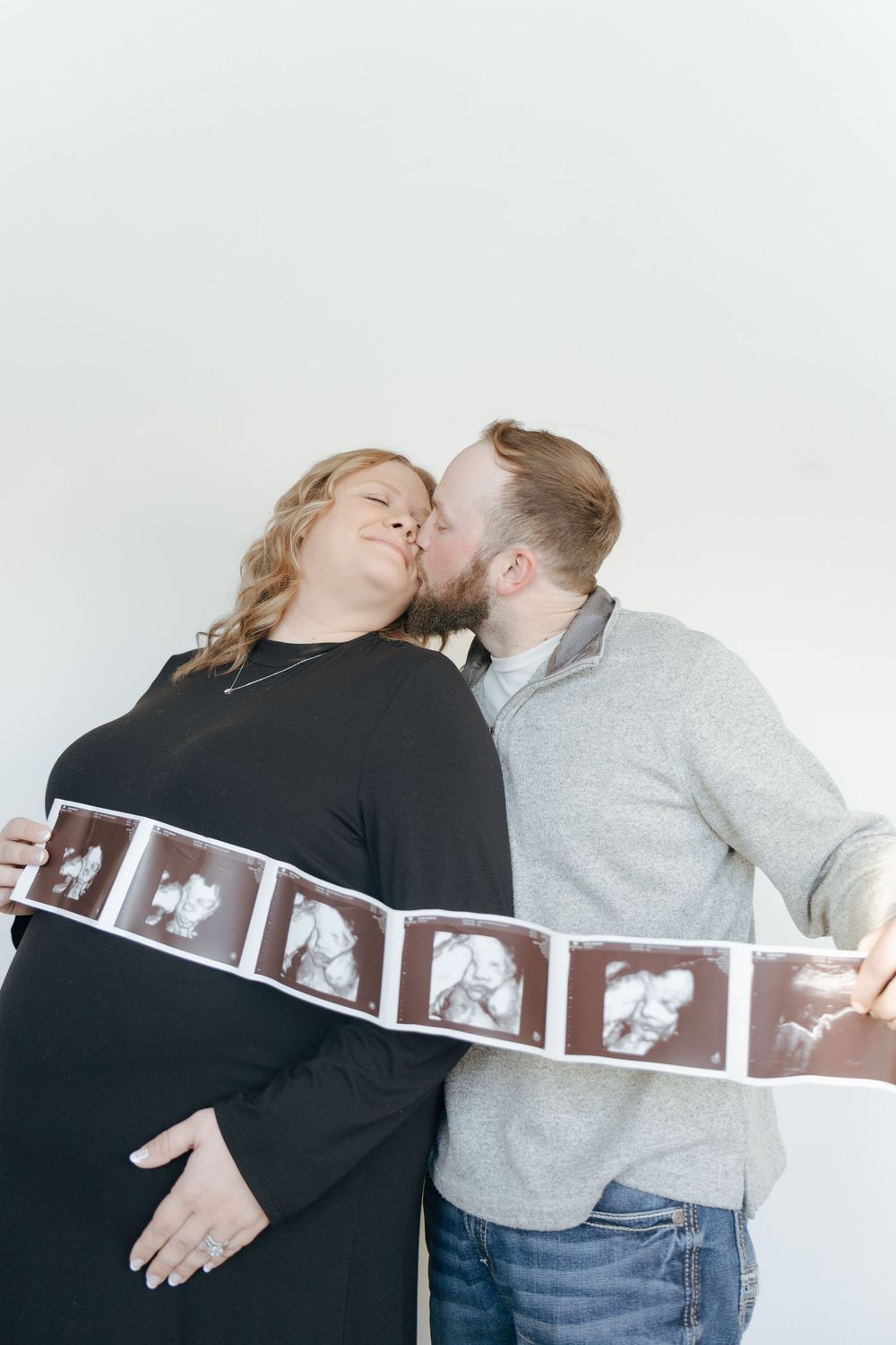 A couple shares a kiss while holding up a strip of ultrasound photos against a white background.