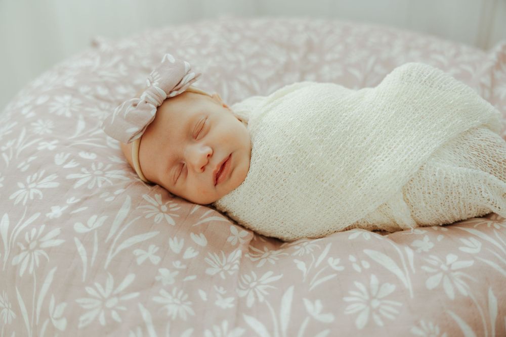 Newborn baby sleeping peacefully on floral patterned bedding while wrapped in a soft white swaddle blanket.