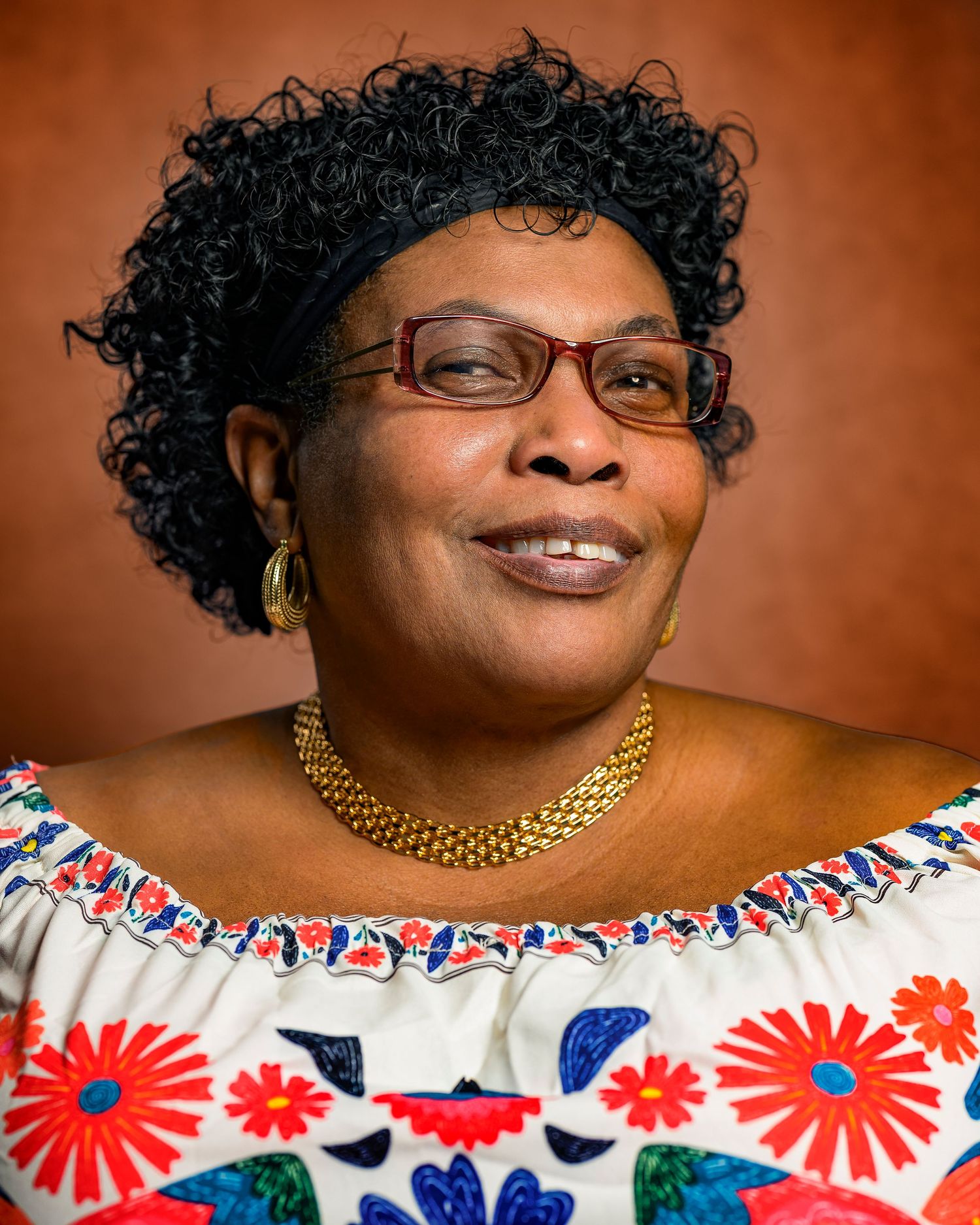 Woman in glasses and floral off-shoulder top wearing gold necklace smiling joyfully.