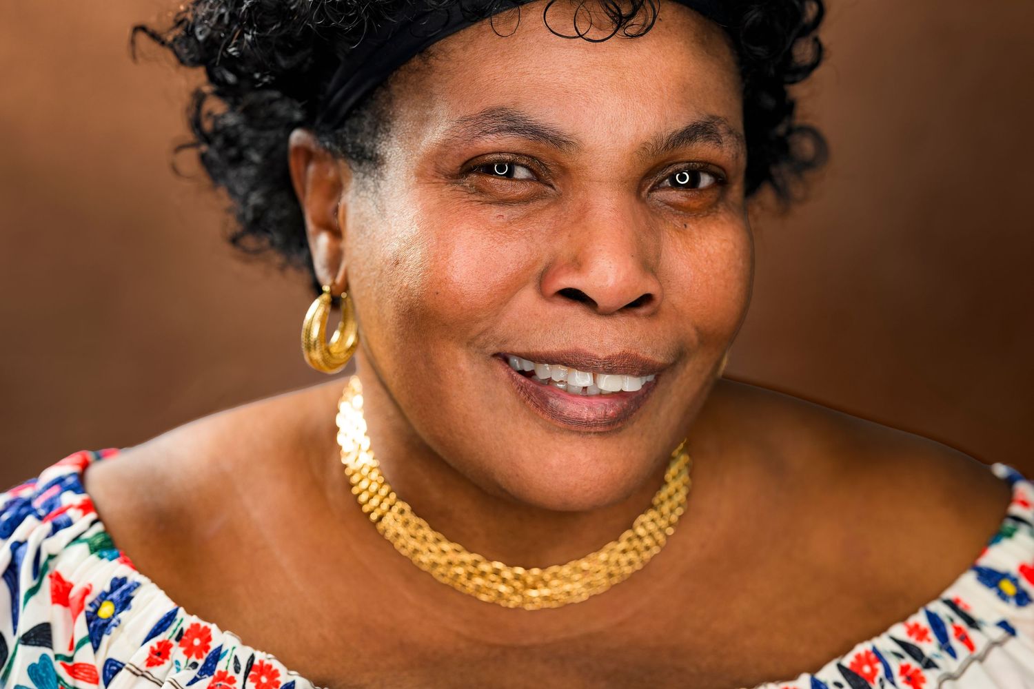 Close-up portrait of smiling woman wearing gold necklace and floral top outdoors.