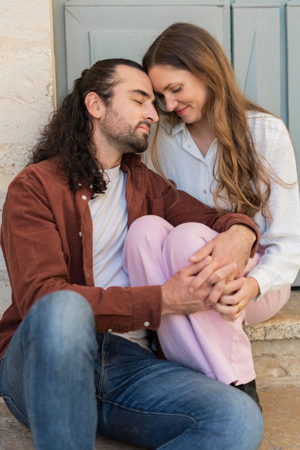 Couple in casual attire sharing an intimate moment while sitting together against a wall with blue shutters.