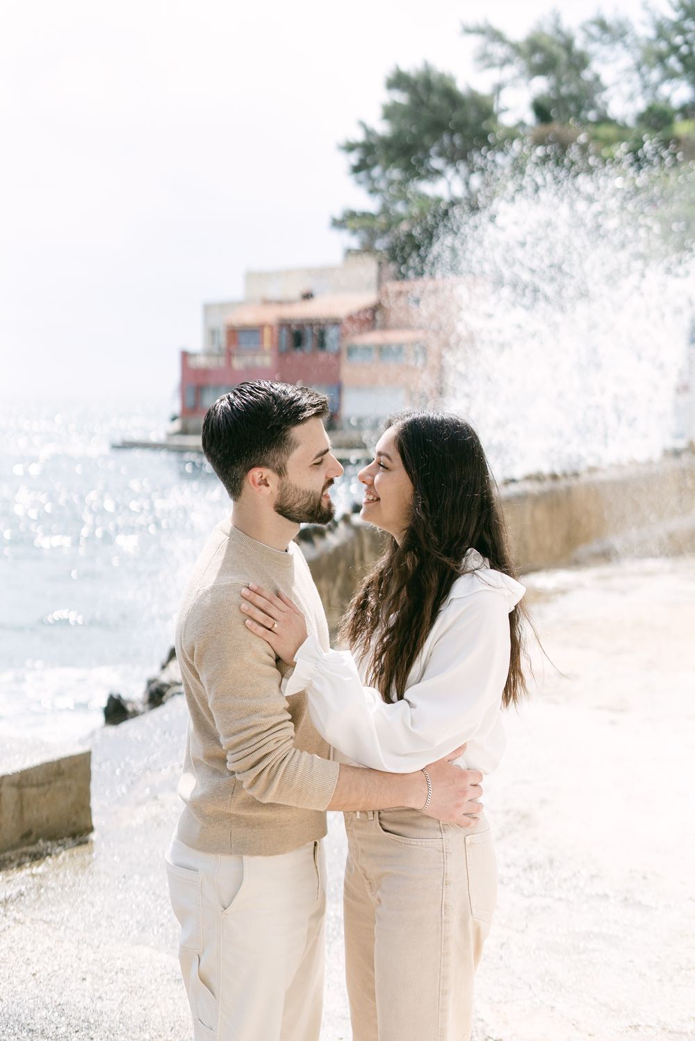 Romantic couple in neutral tones embrace by the ocean as waves splash against rocks on a sunny day.
