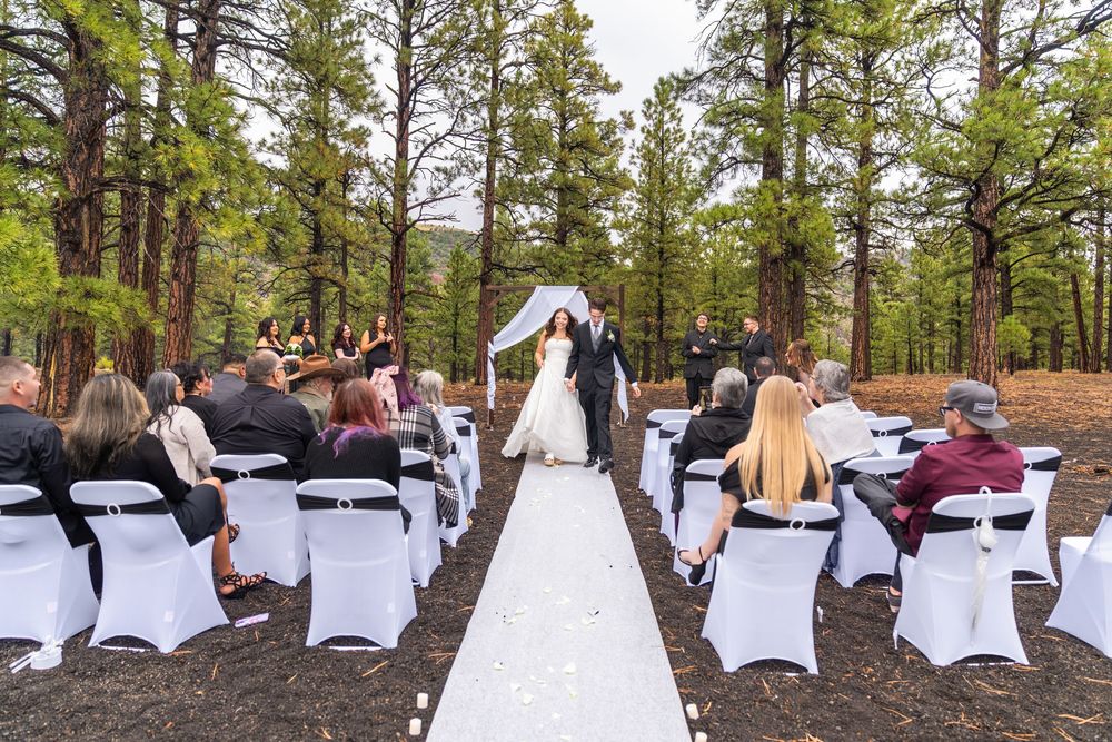 A wedding ceremony in a forest setting with white chair covers and a long white aisle runner.