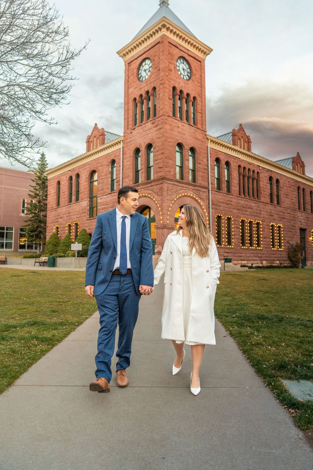 A couple walking together in front of a historic red brick clock tower building.