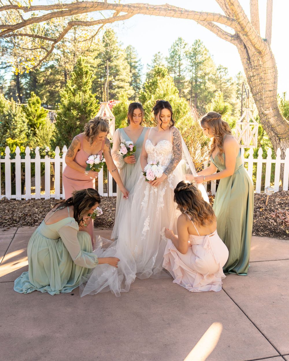Bridesmaids in sage green dresses sit together on a white porch with a picket fence at sunset.